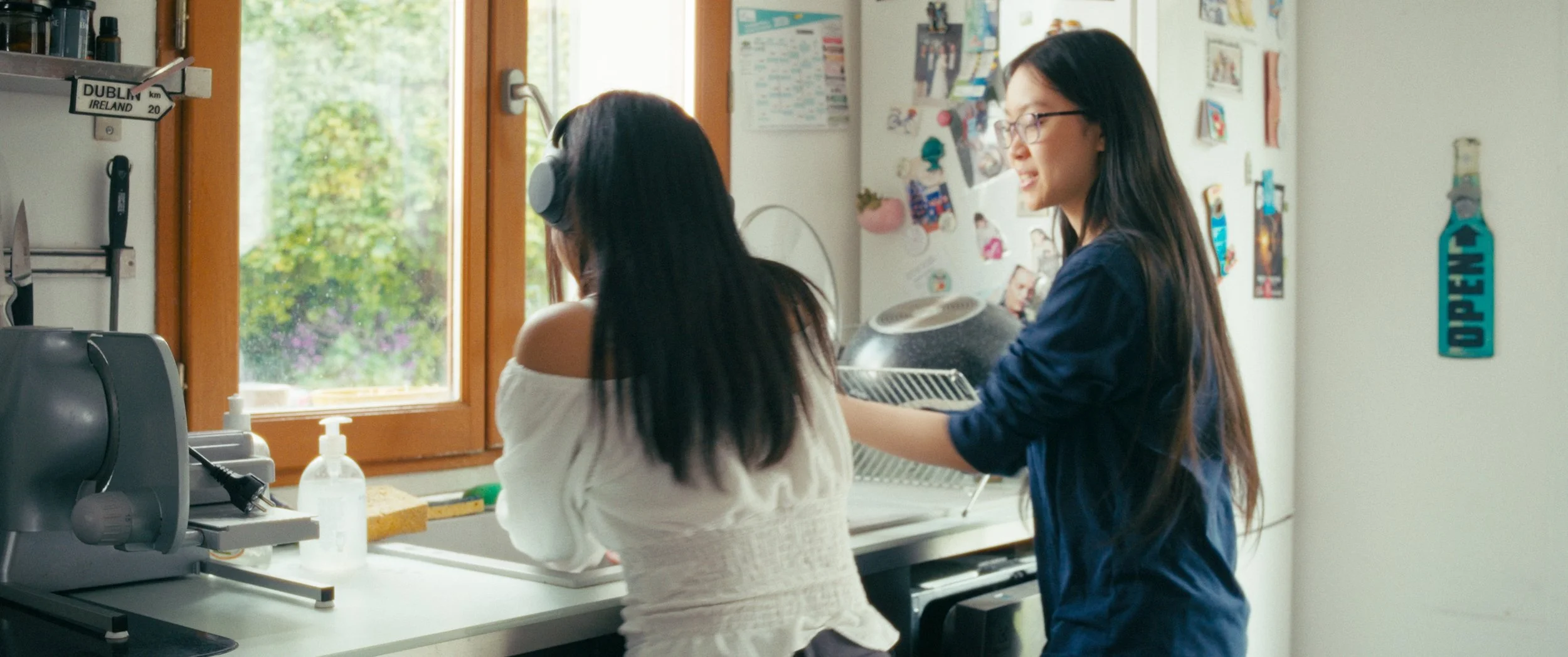 Two women in kitchen, one wearing headphones, washing dishes at sink, the other standing nearby smiling, with colorful photos and posters on wall, window showing greenery outside.