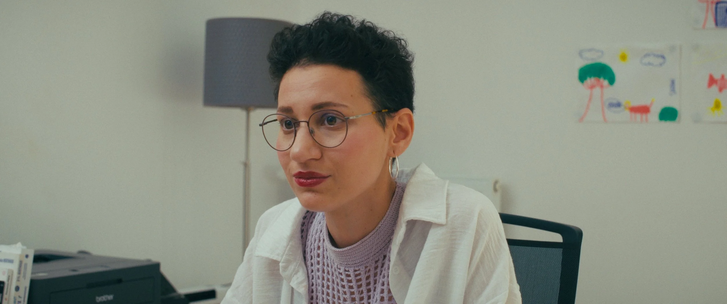 A woman with short dark hair, wearing glasses and hoop earrings, sitting at a desk in an office. Behind her, there is a lamp and a child's drawing on the wall.