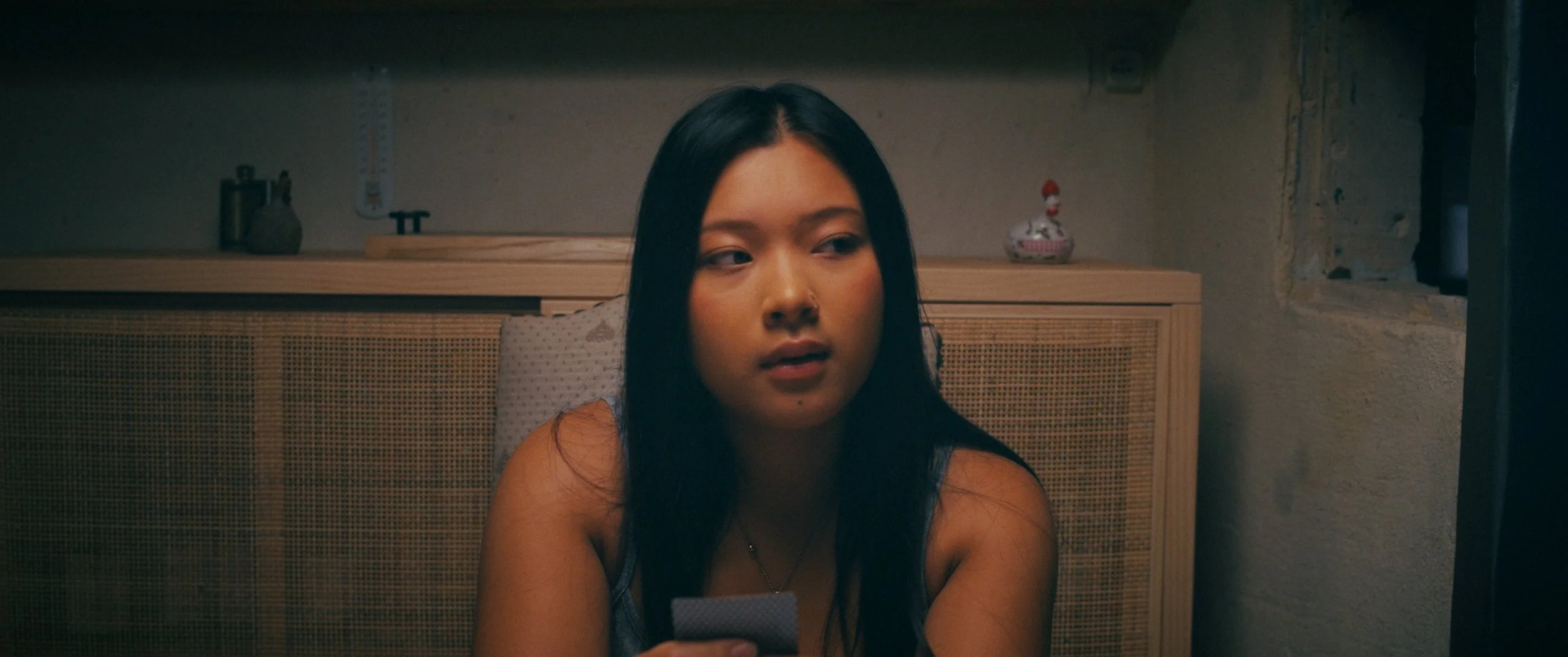 A young woman with long dark hair sitting in a dimly lit room, looking to her left while holding cards in her hand, with a wooden cabinet and decorative objects in the background.