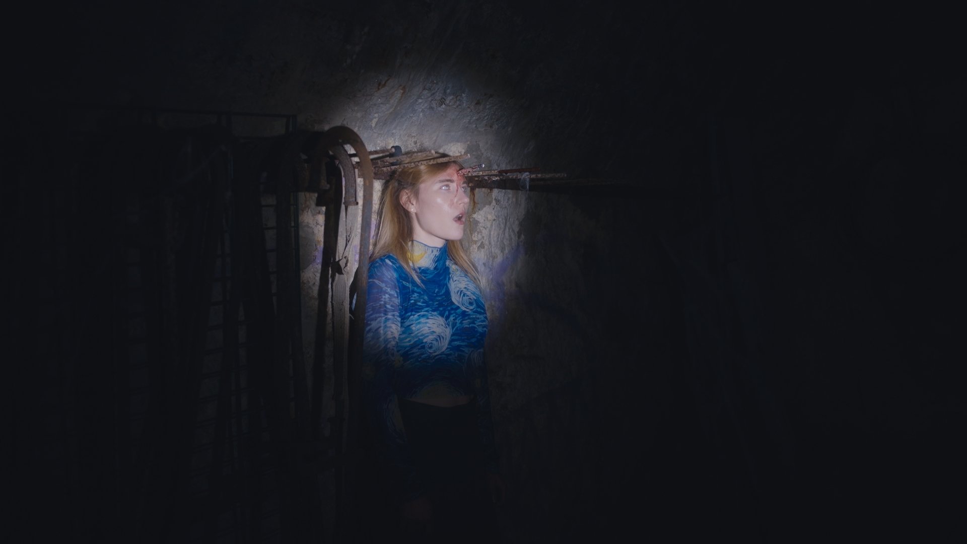 A woman with long blonde hair and painted blue top is shown with her mouth open with a rusty metal rod passing through her head wide shot.