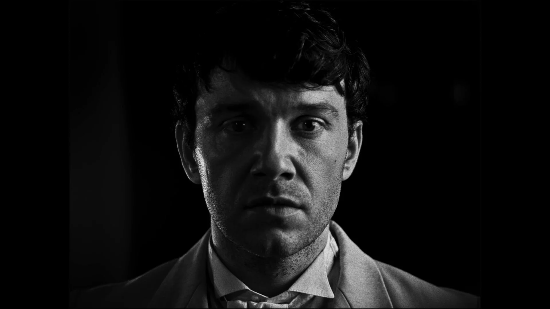 Black and white close-up portrait of a young man with curly dark hair, wearing a suit and tie, against a dark background.