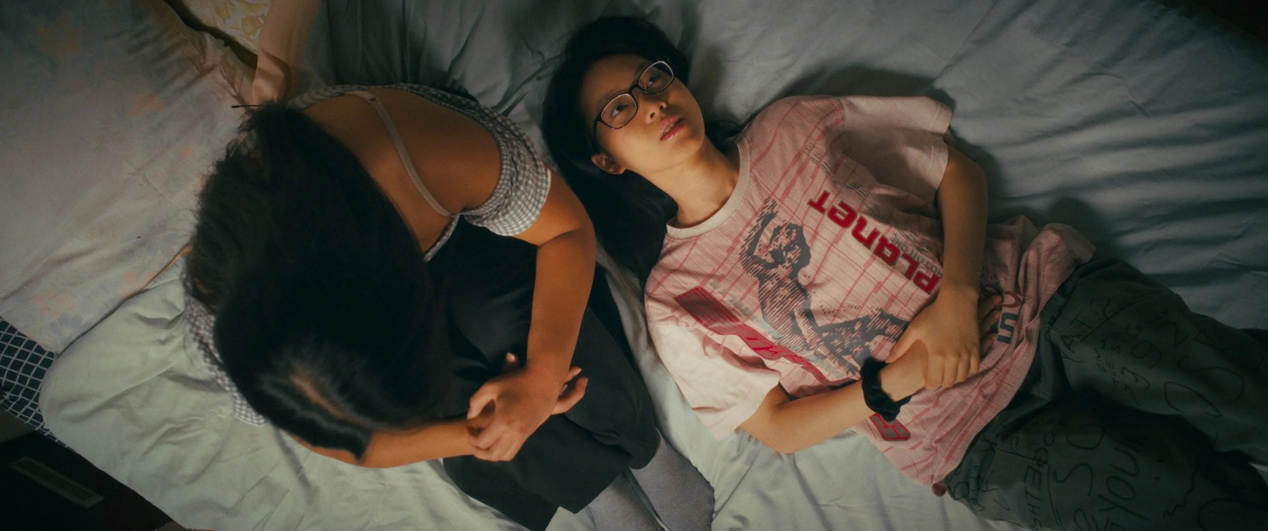 Two girls lying on a bed, one with glasses and long black hair wearing a pink graphic t-shirt, and the other with long black hair in a ponytail wearing a black and white shirt. The girl with glasses is looking at the ceiling.