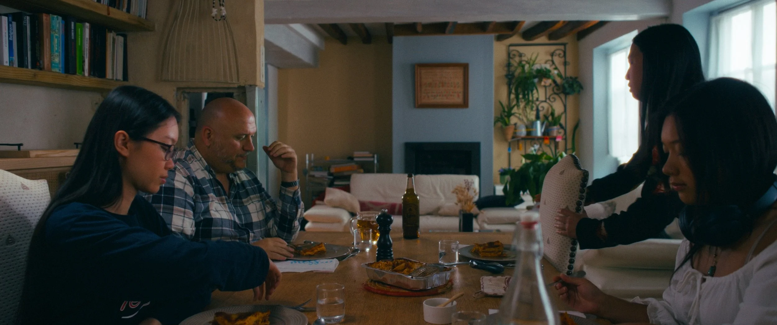 A group of five people sitting at a wooden dining table in a cozy, well-lit room with plants and books, having a meal with pizza, drinks, and condiments.