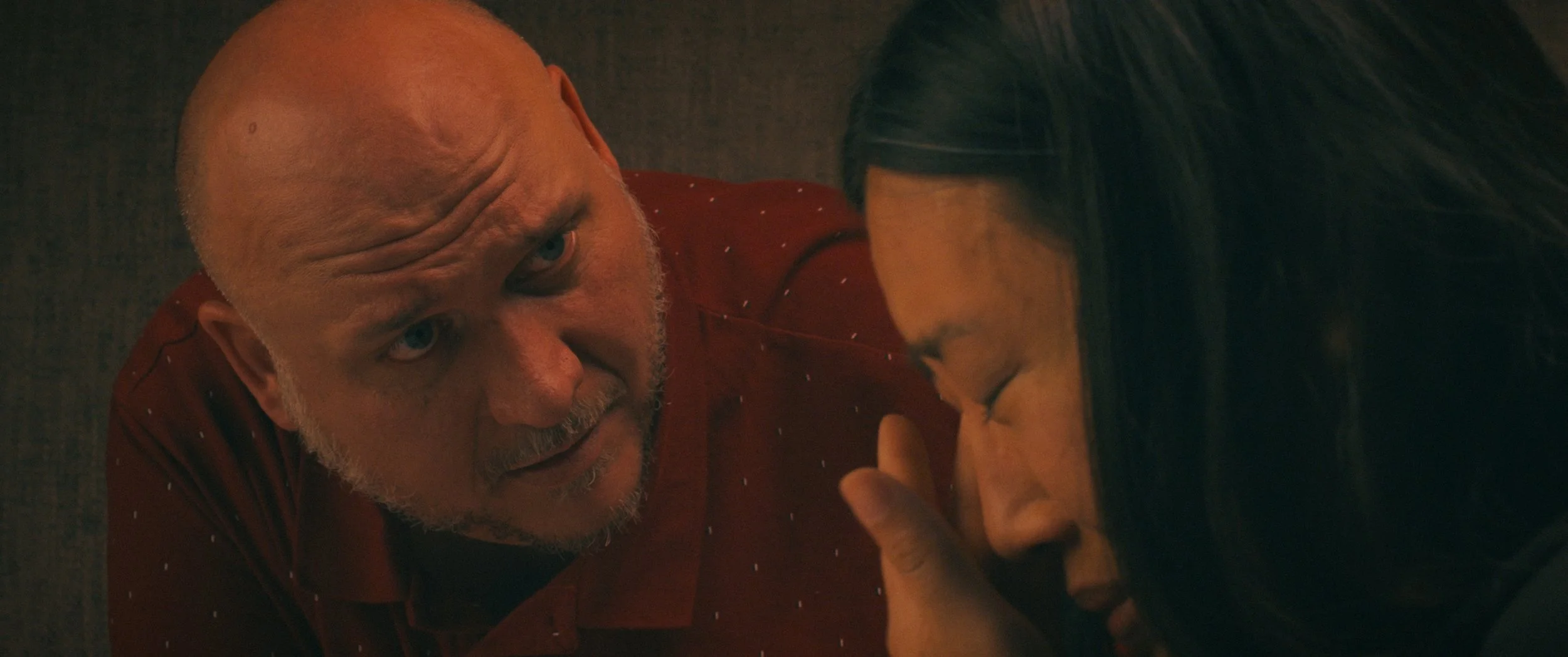 An elderly man with a bald head and gray beard leans over a woman with long dark hair, engaged in an intense emotional moment.