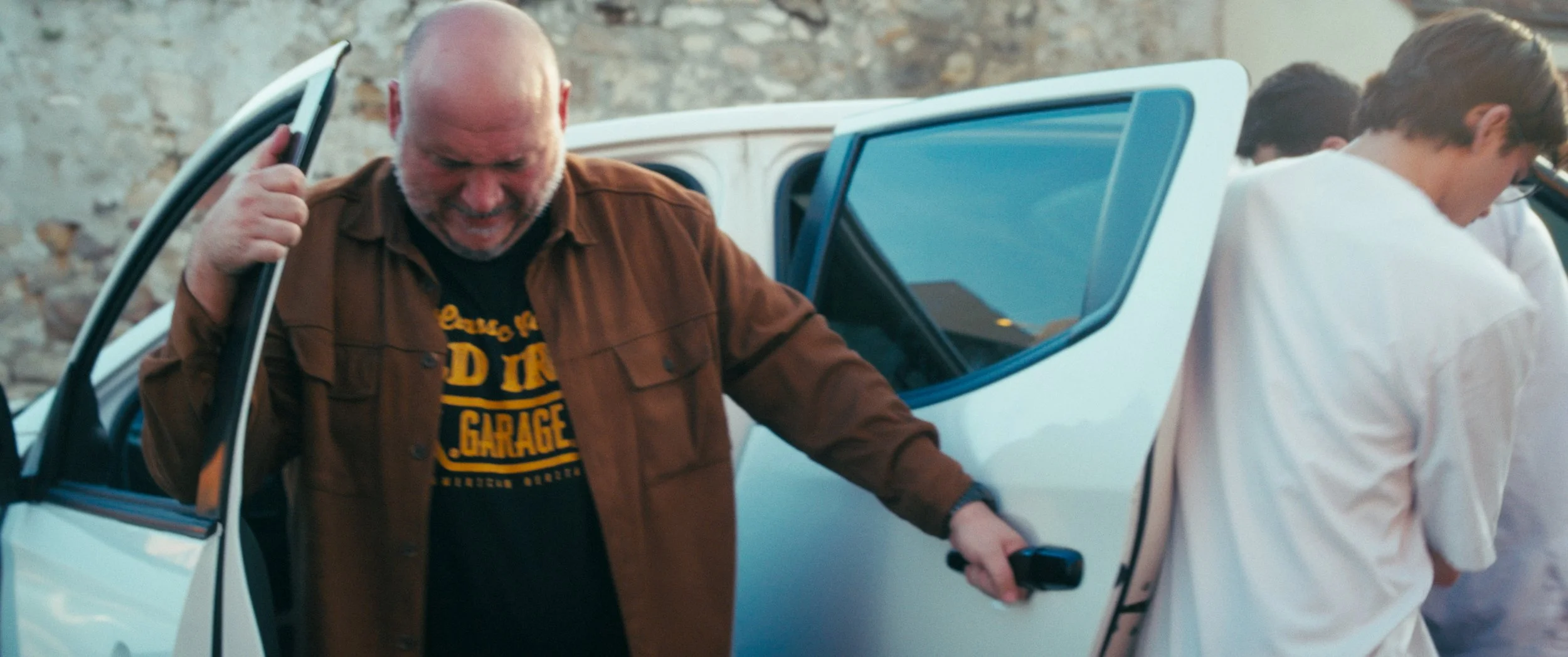 A man with a bald head and gray beard stepping out of a white pickup truck, smiling, wearing a brown jacket and black t-shirt with yellow text, with two younger men in white shirts standing nearby in front of a stone wall.