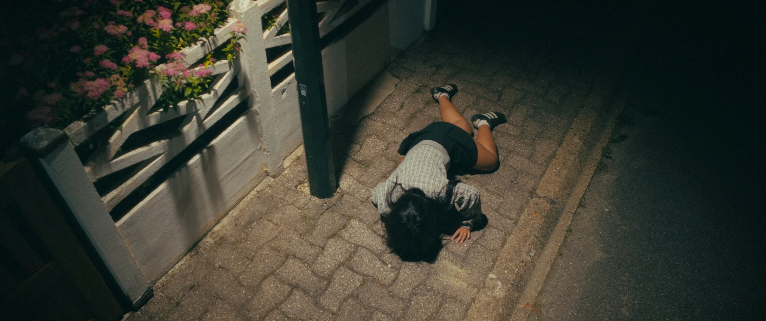 A girl lying on the sidewalk at night, wearing light blue jacket, near a flower bed with pink flowers, beside a fence and a lamp post.
