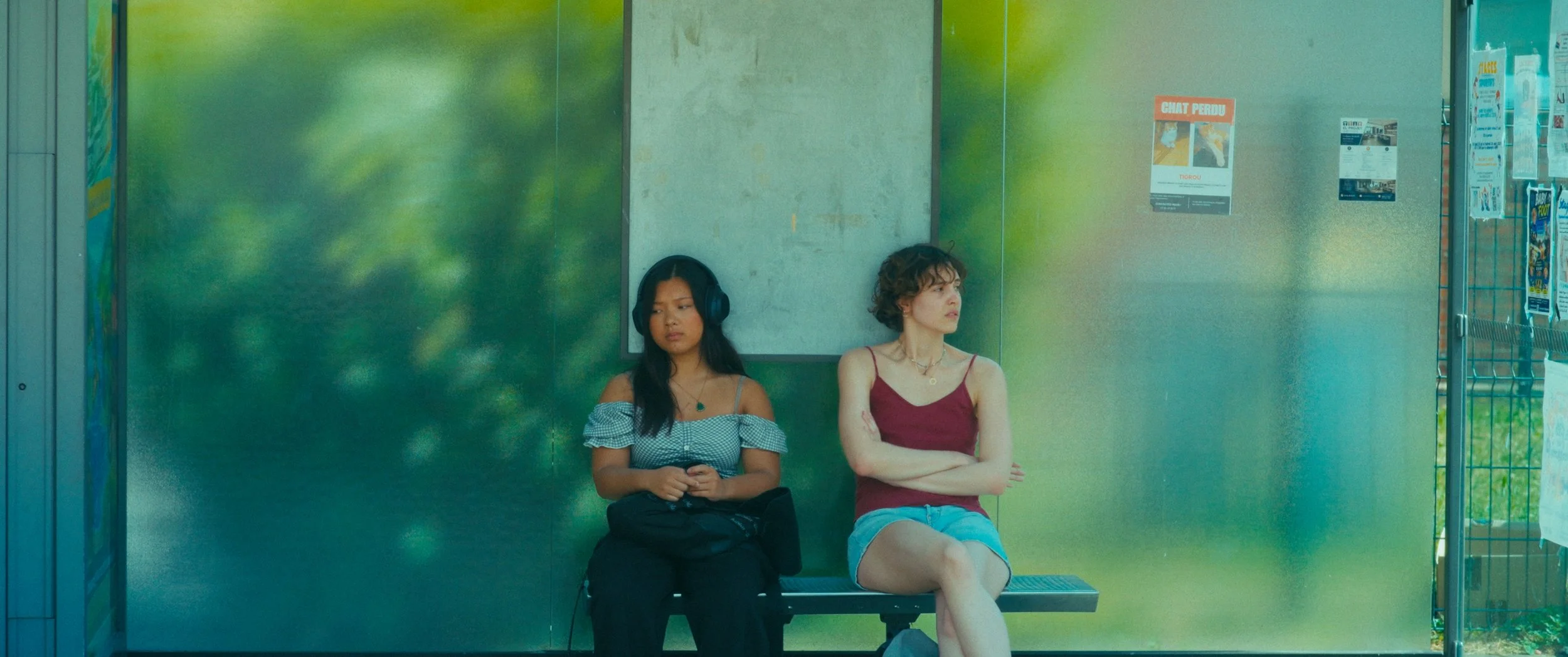 Two women seated at a bus stop, one with headphones and the other with folded arms, in a shaded waiting area with advertisements on the wall.