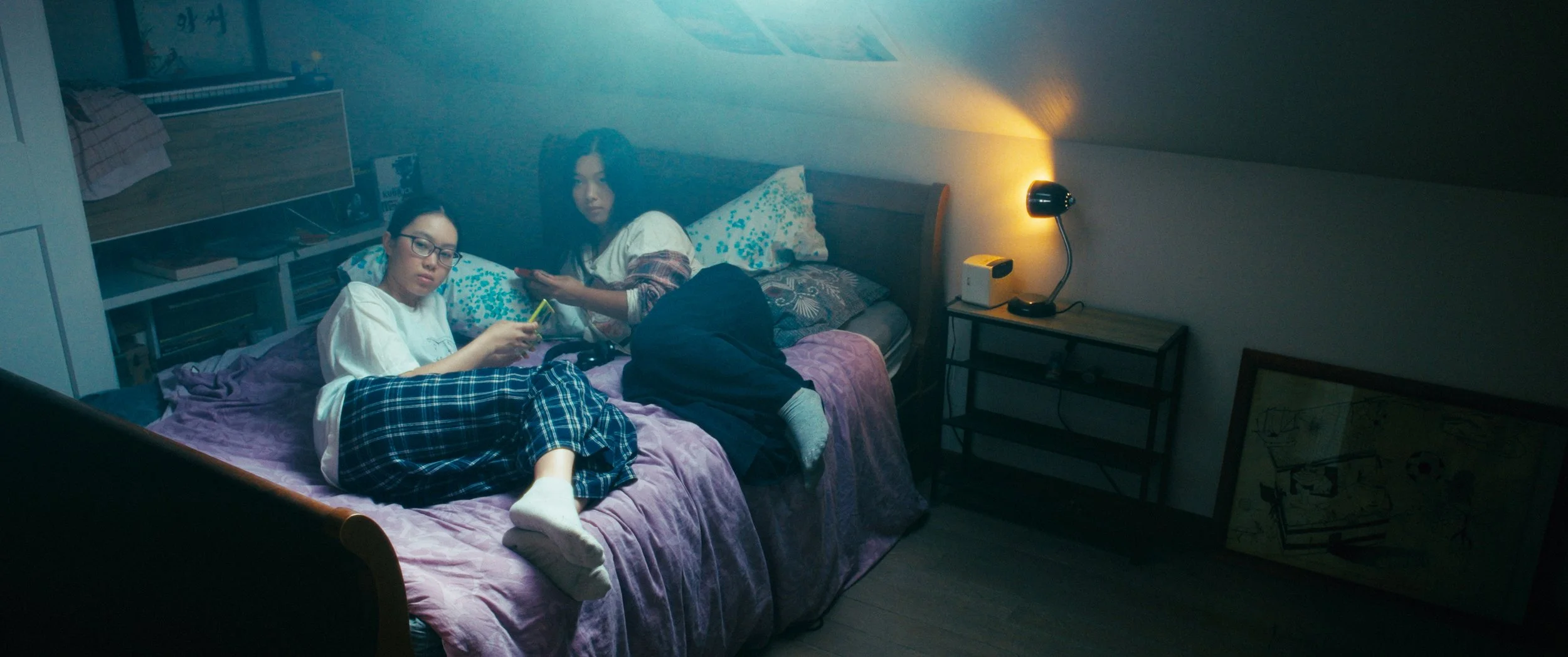Two girls sitting on a bed in a dimly lit bedroom, one using a smartphone and the other holding a remote, with a bedside lamp on and a small shelf nearby.
