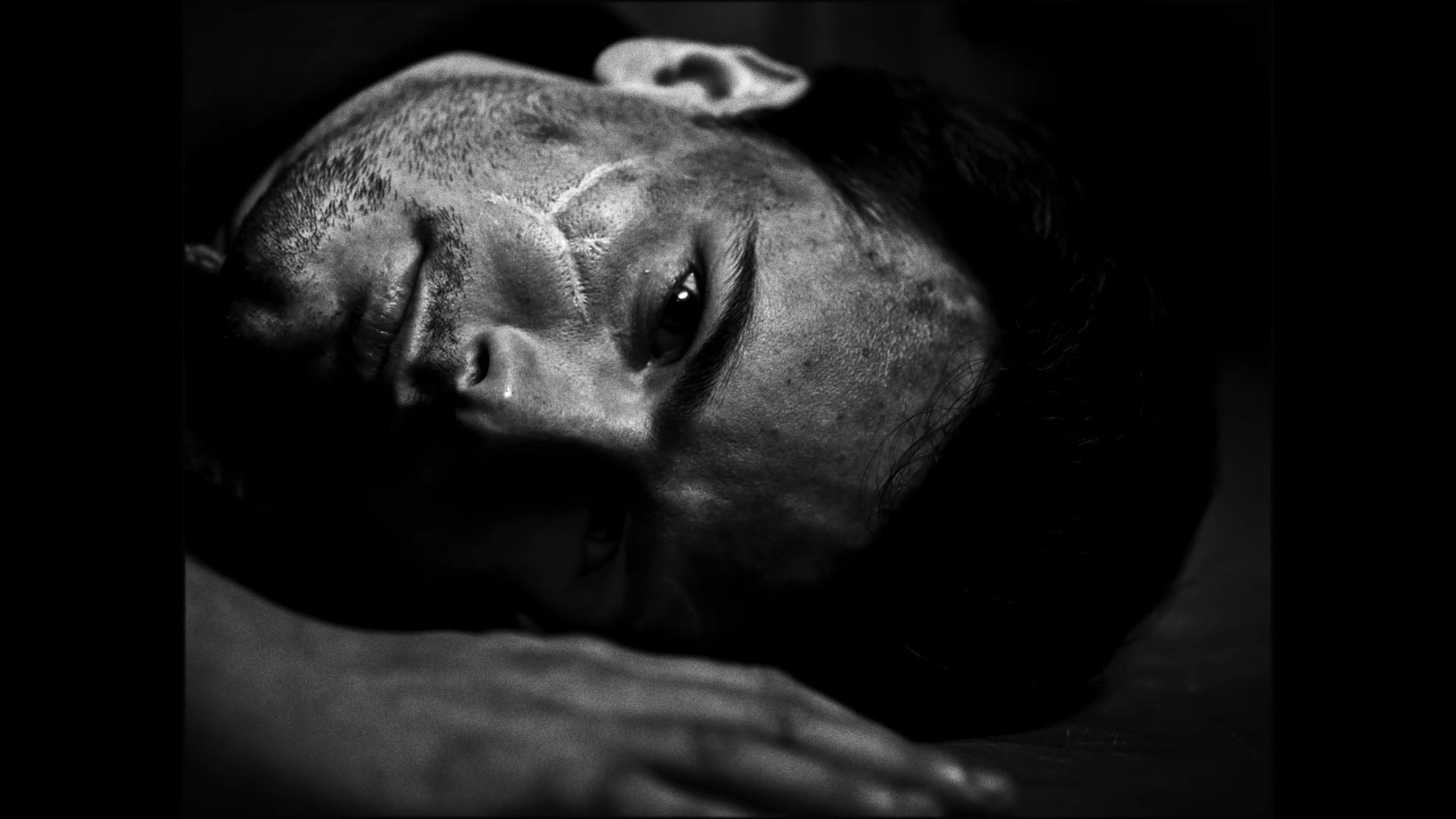 Black and white photo of a young man lying down with head turned to the side, eyes slightly open, showing facial details and short hair.