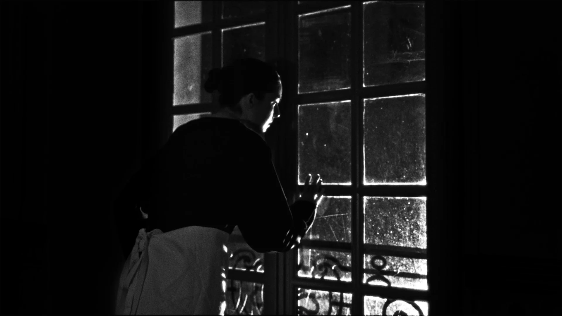 A person standing near a window, looking outside in black and white. The person has dark hair tied back and is wearing a dark top, with their hand resting on the window pane.