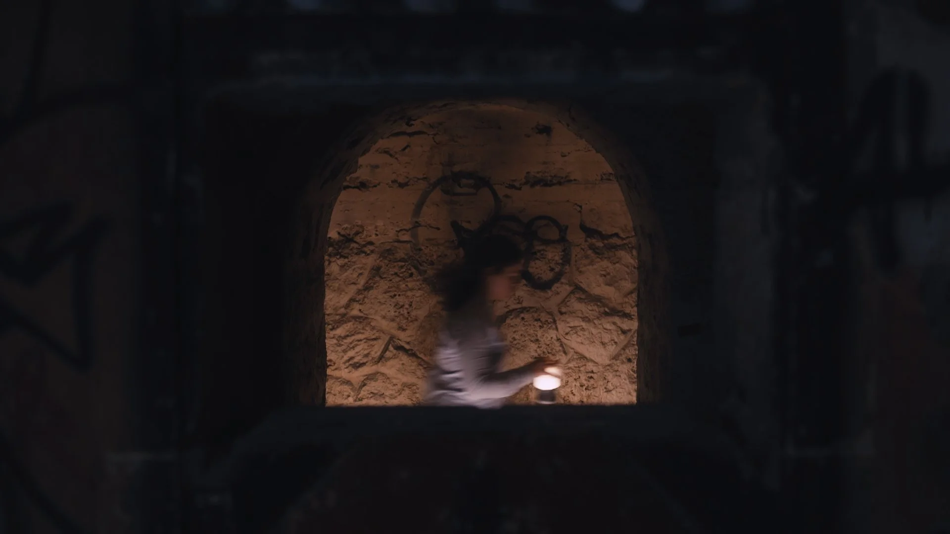 A person walking through a dark tunnel with stone walls, holding a small light or candle, with graffiti on the stone wall behind them.