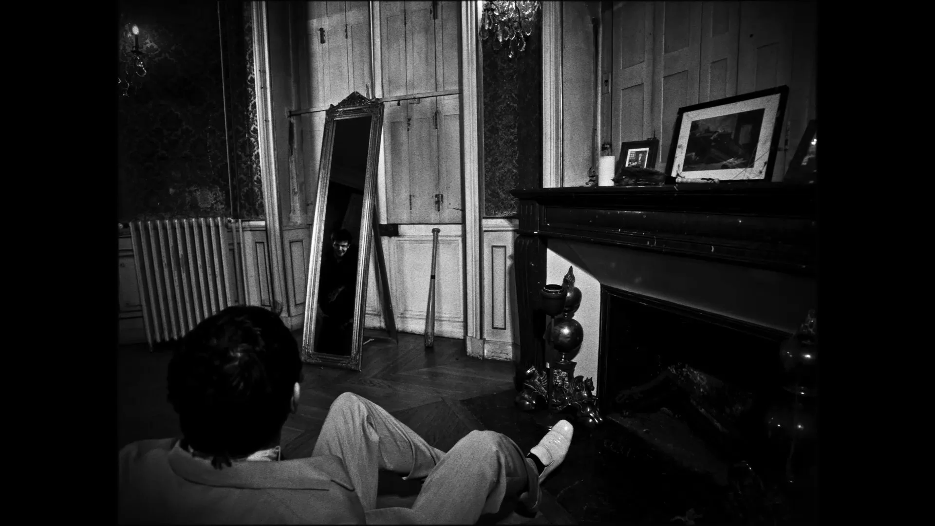 A man sitting on the ground, ornate mirror leaning against a wall in a dimly lit with the reflection showing another person, vintage-style room. The room has wooden paneling, a fireplace, framed pictures on top.