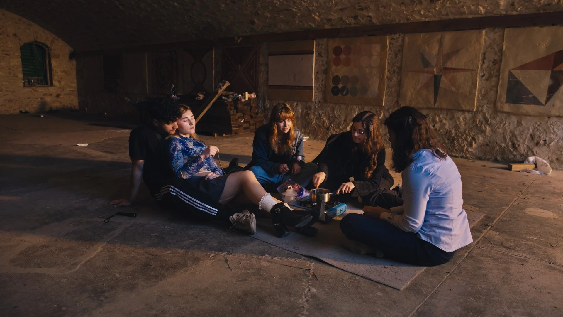 Group of teenagers sitting on a mat in a rustic, stone-walled room, gathered around a small portable stove with cooking utensils and snacks, engaging in conversation.