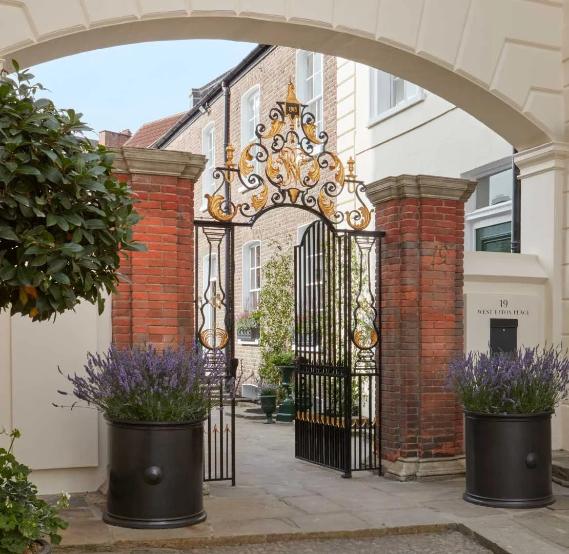 View of an ornate wrought iron gate with gold accents, flanked by brick pillars, leading into a courtyard with potted lavender plants and a building in the background.