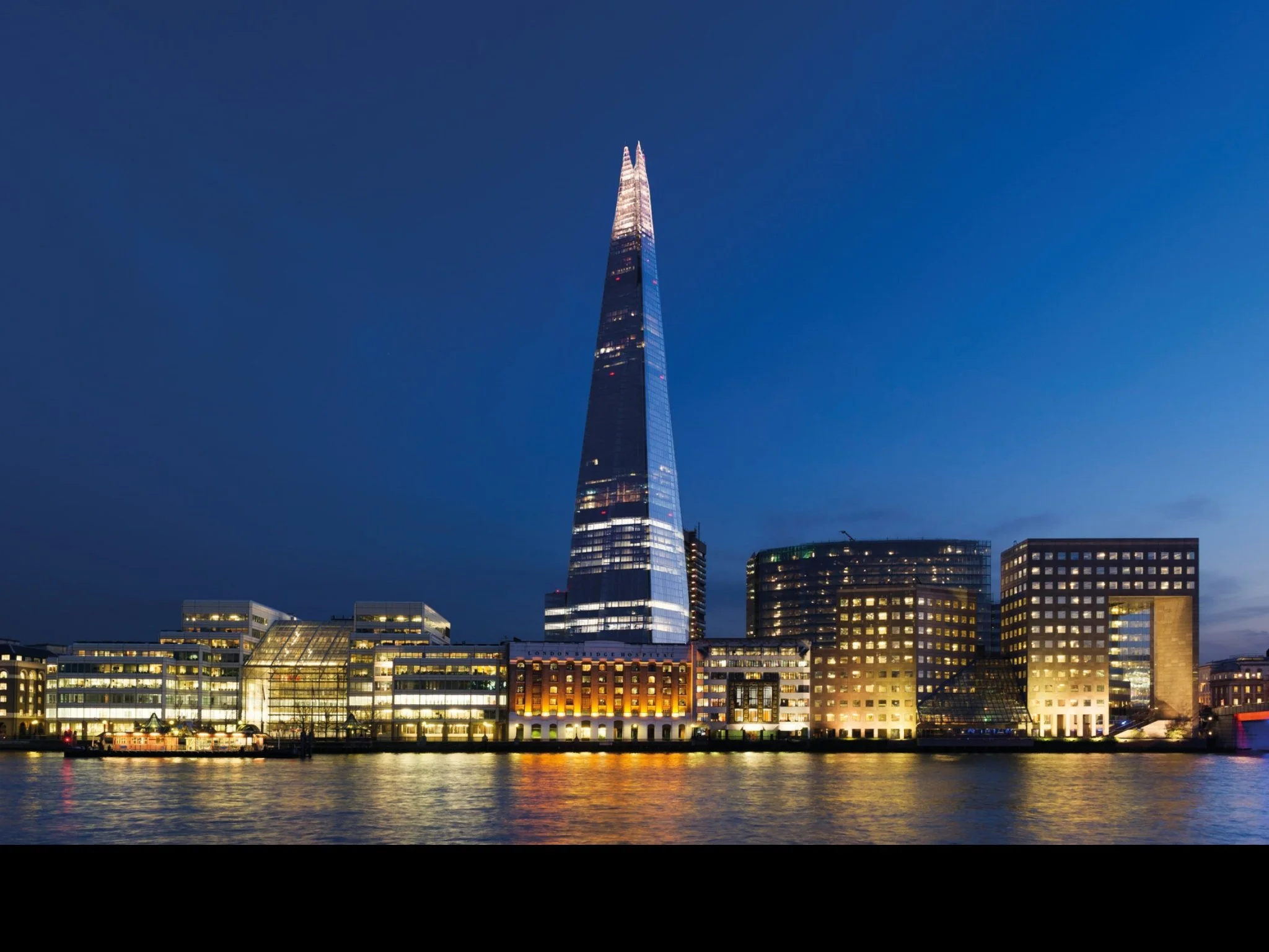Night view of the London skyline with The Shard illuminated, reflecting on the River Thames, and surrounding modern buildings with lit windows.