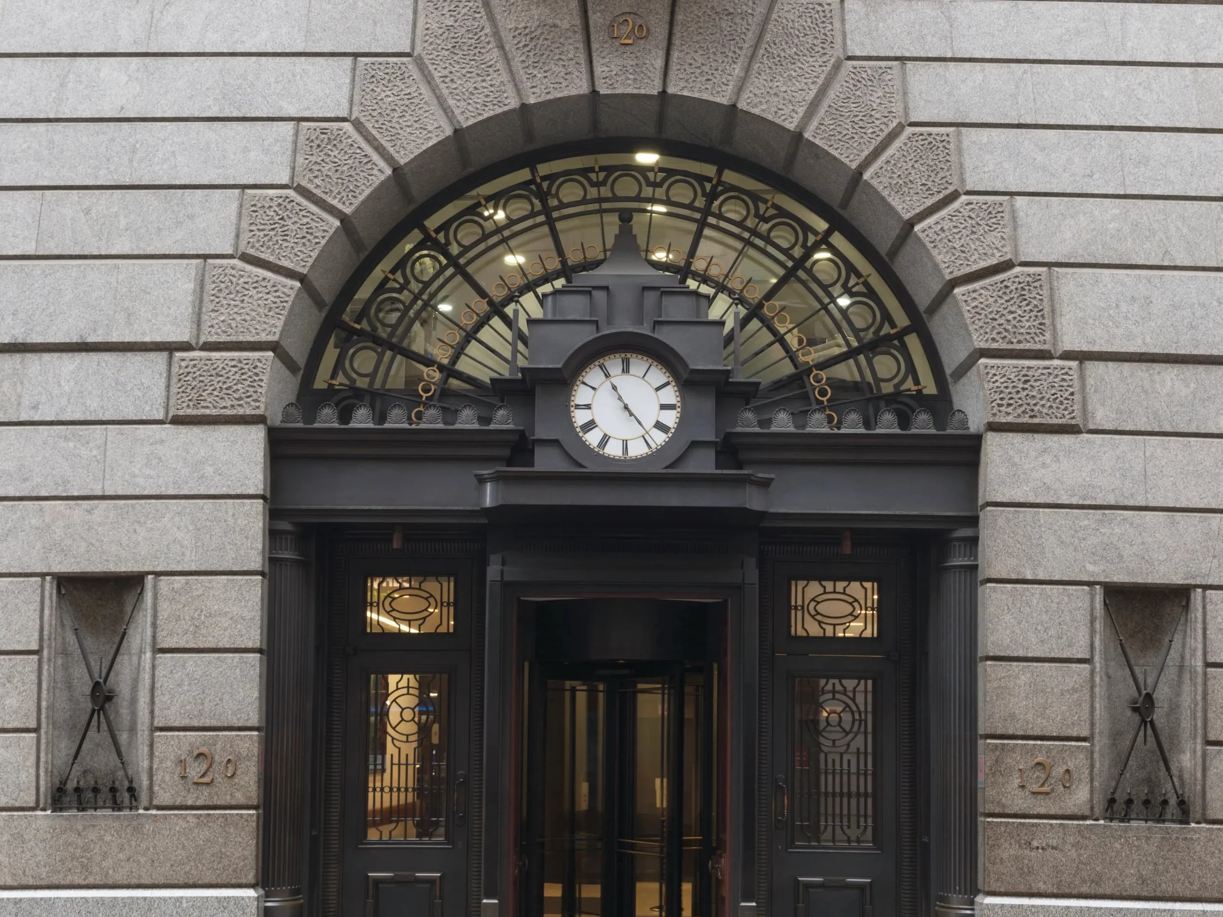 Entrance of a historic building with a large, arched clock above the door showing the time as 4:32, surrounded by ornate metalwork, with the building's address number 120 displayed on the stone facade.