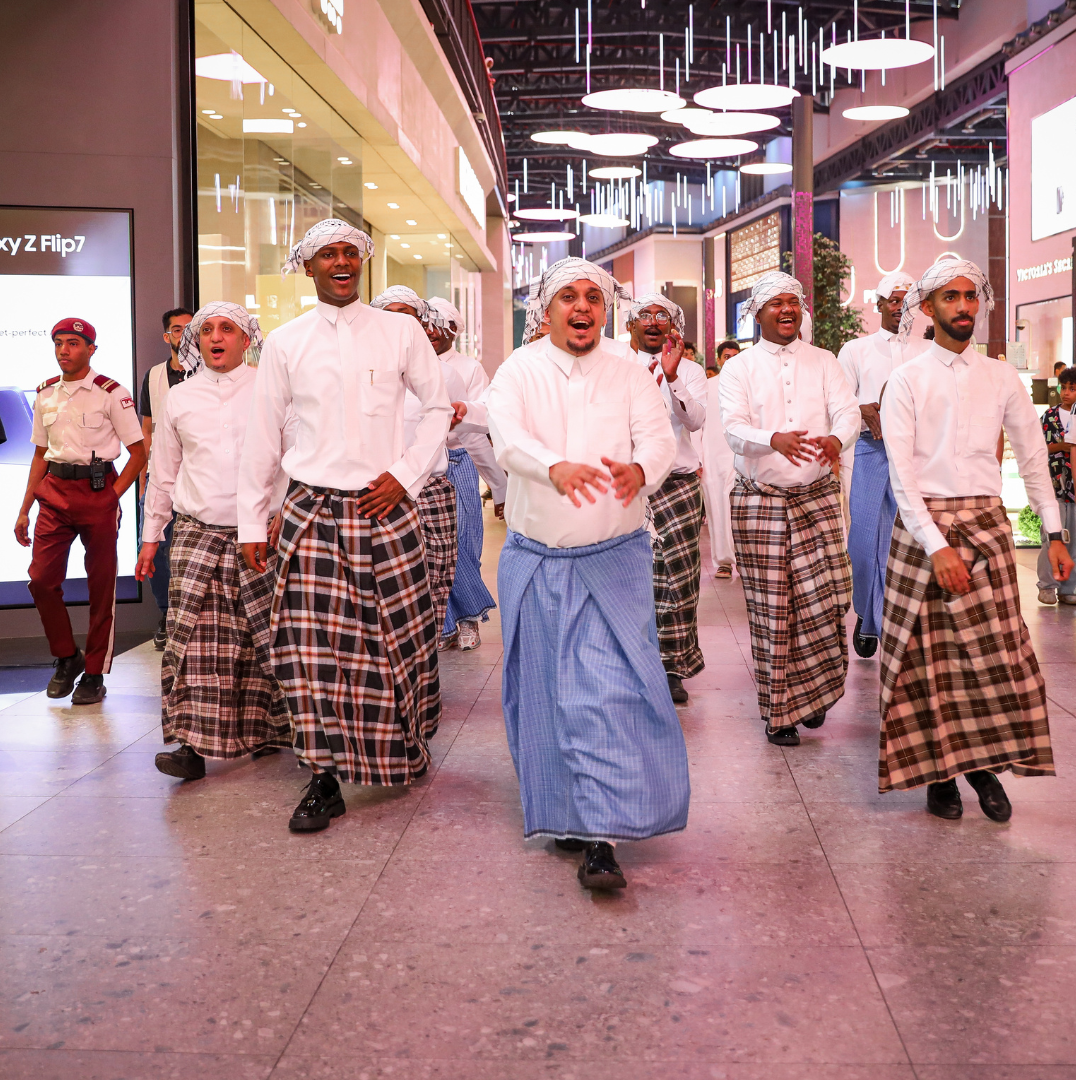Group of men in traditional Middle Eastern attire, including white shirts, checkered sarongs, and head coverings, walking through a shopping mall, smiling and casually talking.