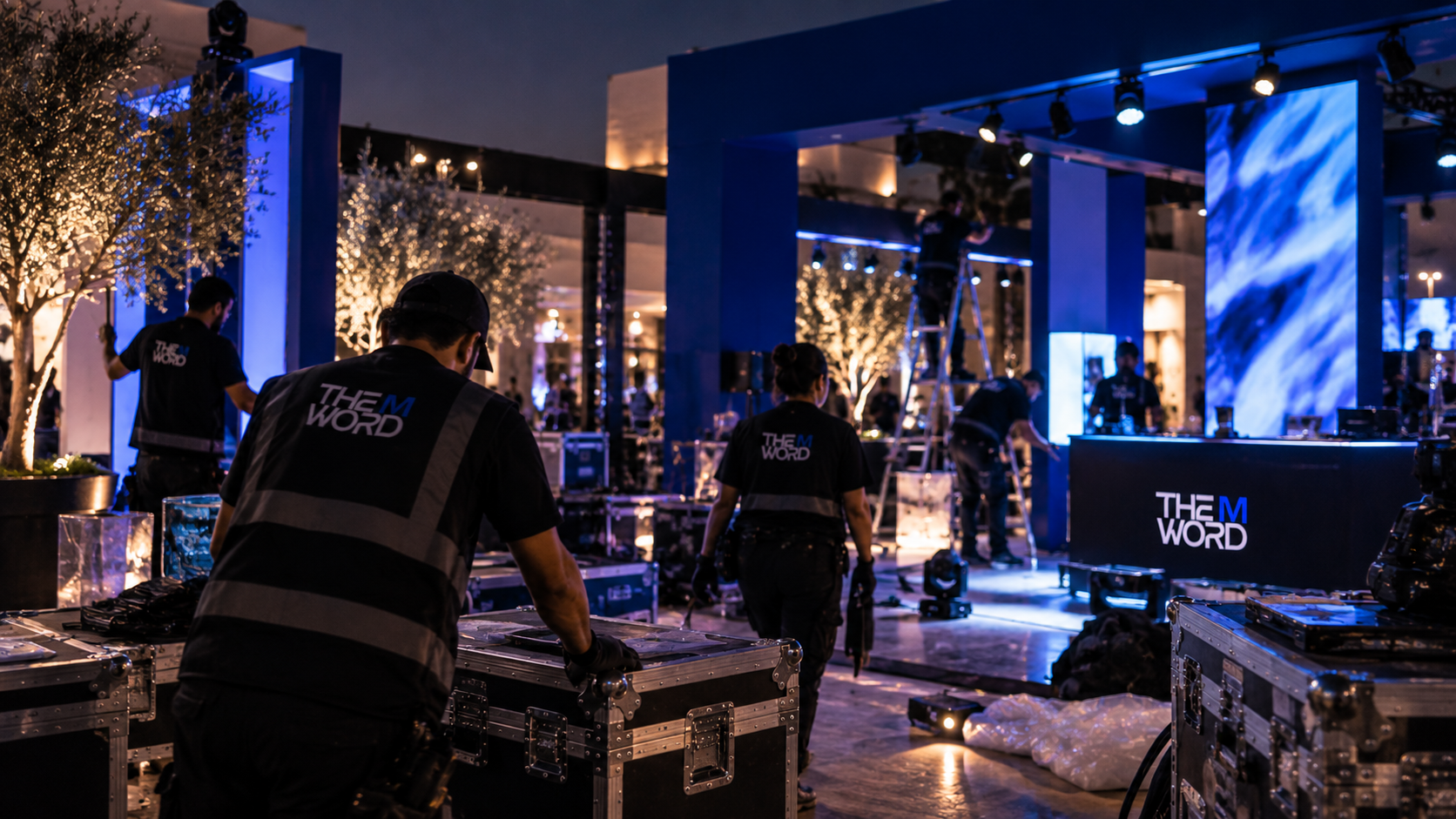 Staff setting up equipment at an indoor event with decorative trees and blue lighting, including a large screen and a DJ booth labeled 'THE M WORD'.