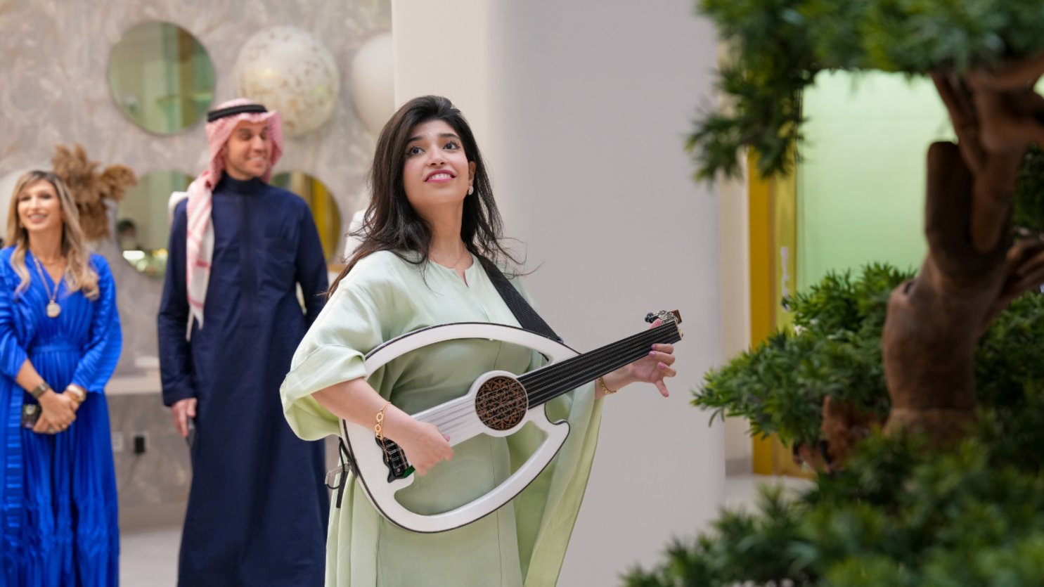 A woman playing a white guitar in a shopping mall, with people watching her and a tree nearby.