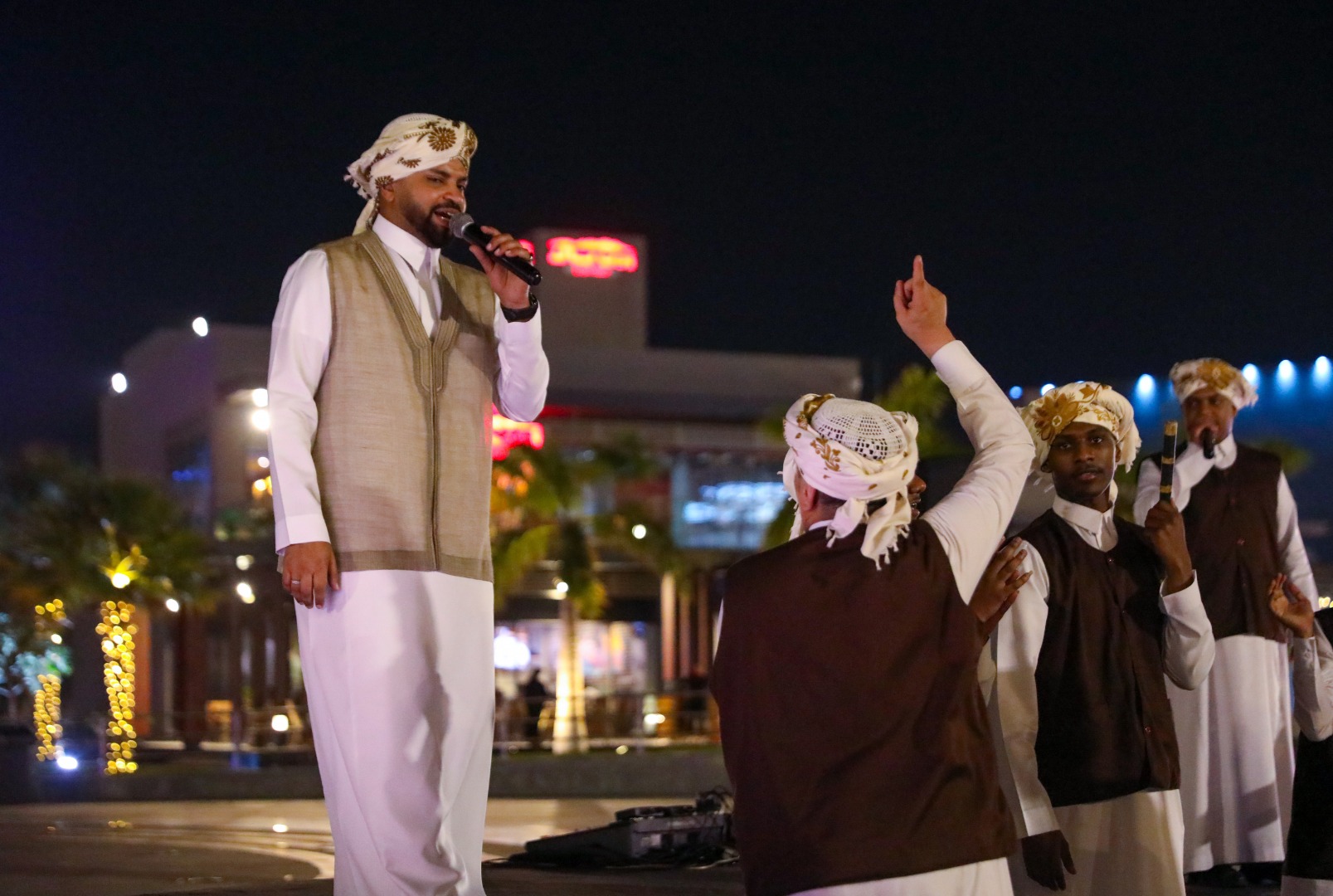 Man in traditional Middle Eastern clothing singing into a microphone at night, with other men in traditional attire, one raising his hand, in an outdoor setting with illuminated trees and buildings in the background.