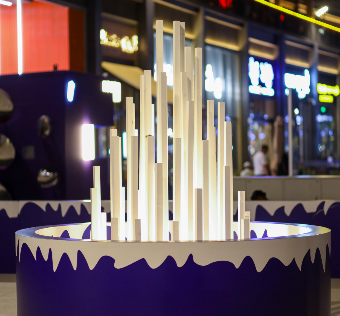 Illuminated modern art sculpture with vertical white light tubes inside a circular dark purple base, located in a shopping mall or entertainment complex at night.