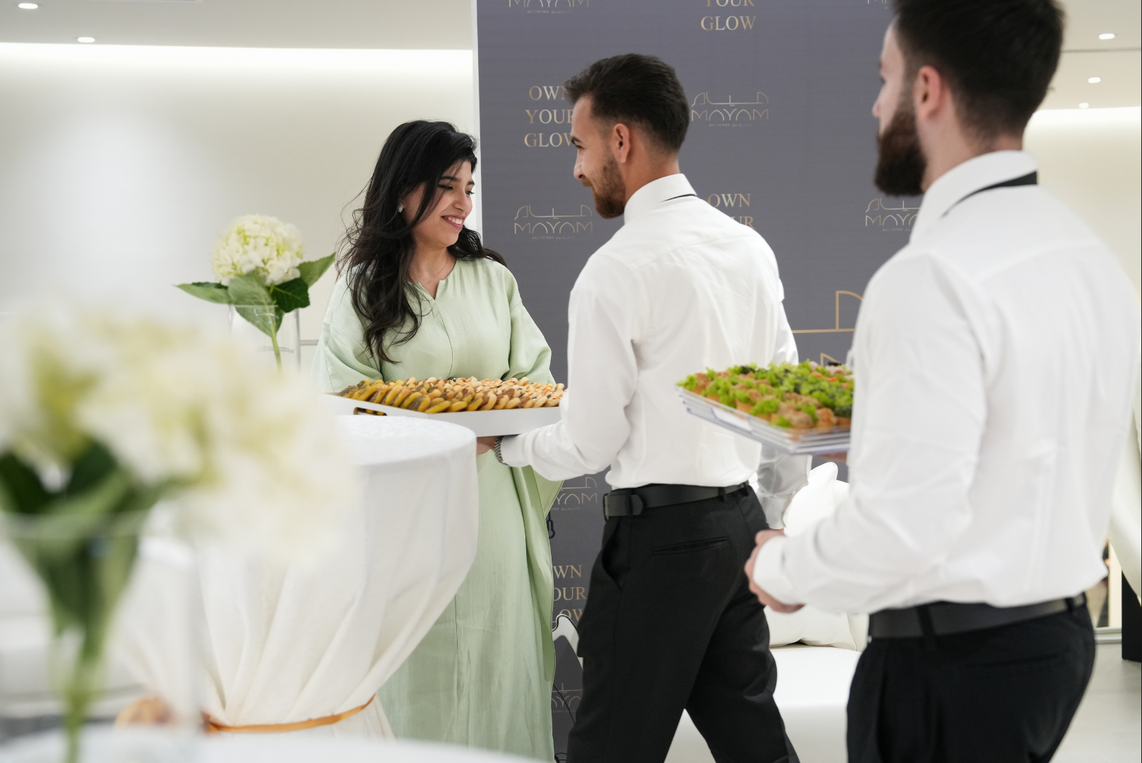 A woman at a reception table receiving food from two waiters. The reception area has a dark backdrop with gold text and white flowers are visible in the foreground.