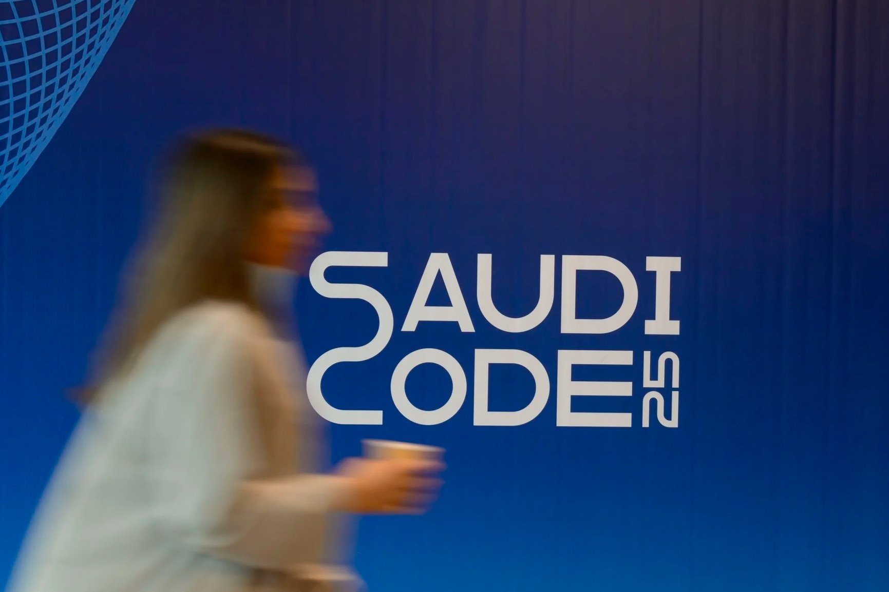 Blurry woman walking past a large blue wall with the text 'Saudi Code 25' printed on it.