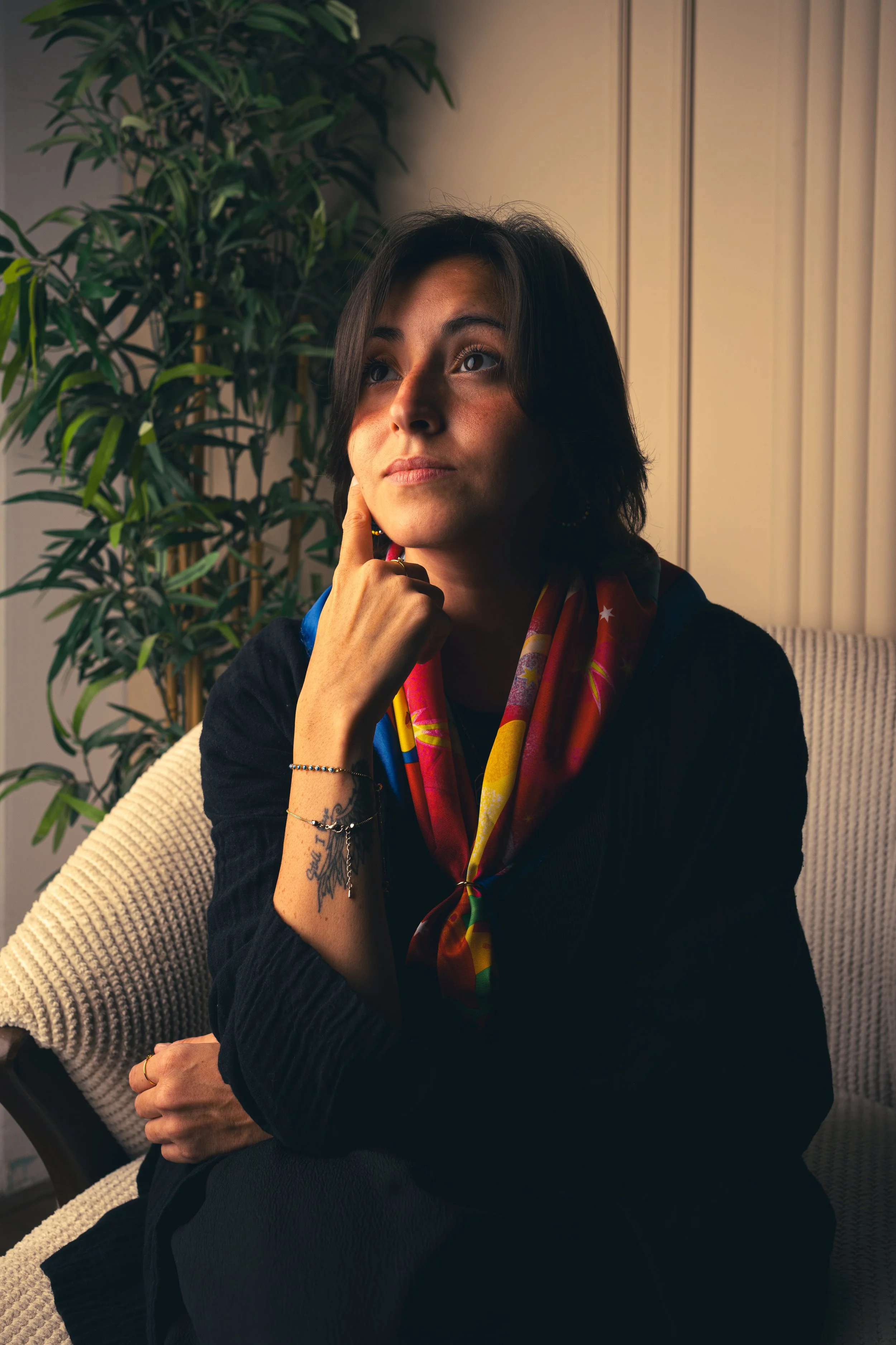 A woman with dark hair, wearing a colorful scarf and black clothing, sitting on a beige textured chair next to a large green plant, with a thoughtful expression, indoor lighting, cream-colored curtains in the background.