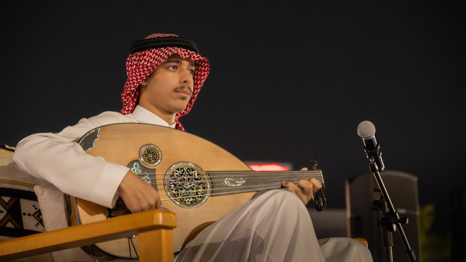 Young man in traditional Middle Eastern attire playing an oud guitar at night with a microphone nearby and a dark sky in the background.