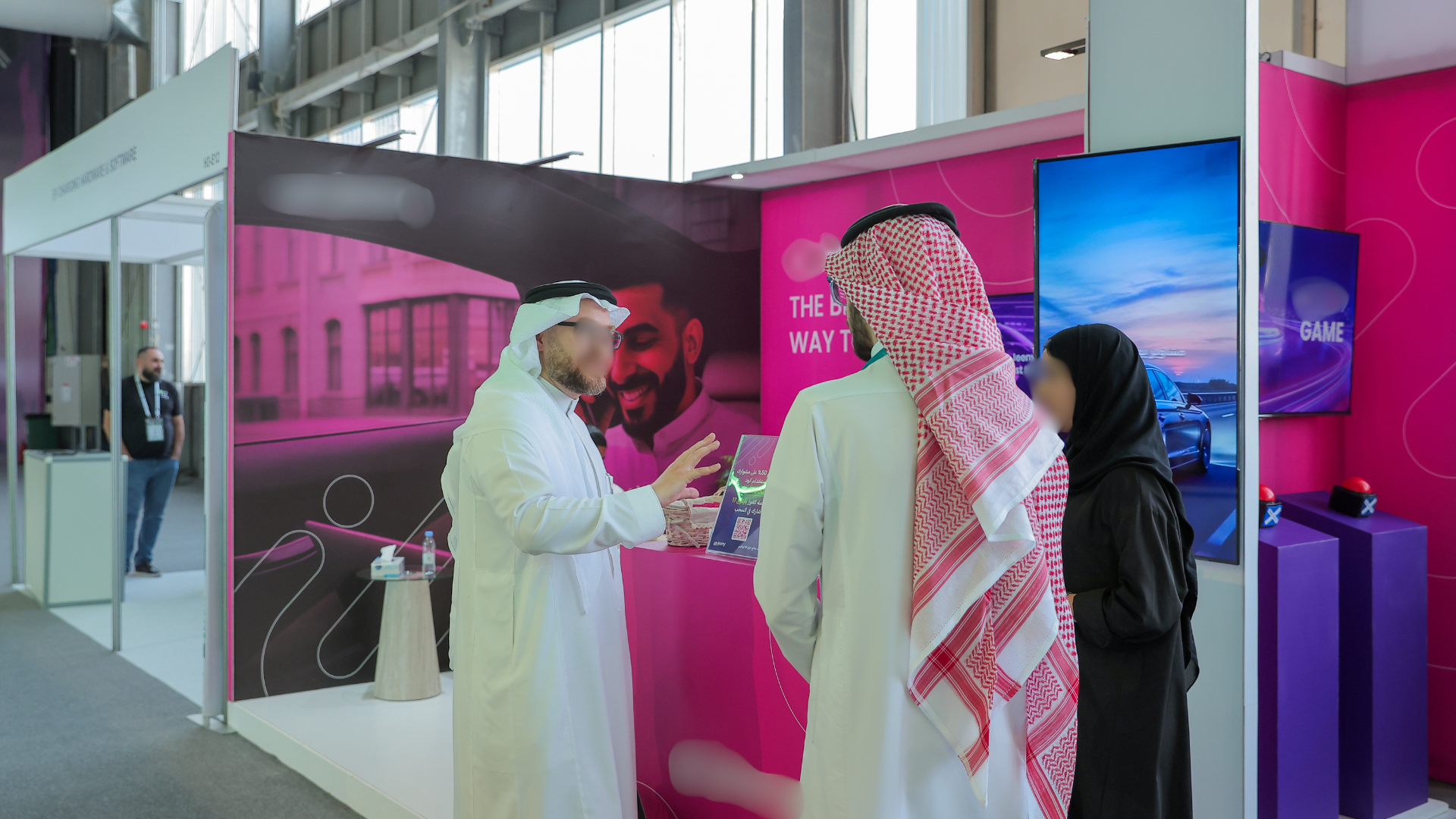People at a trade show booth, talking to a representative, with television screens and promotional material in a brightly colored pink and purple exhibition space.