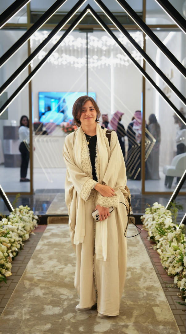 Woman in traditional cream-colored abaya holding a small camera, standing on a floral aisle runner at the entrance of a decorated venue with black geometric neon lighting and glass doors