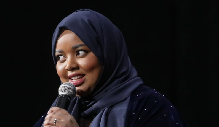 Close-up of a woman wearing a navy blue hijab and dark blue dress, speaking into a microphone on stage against a black background.