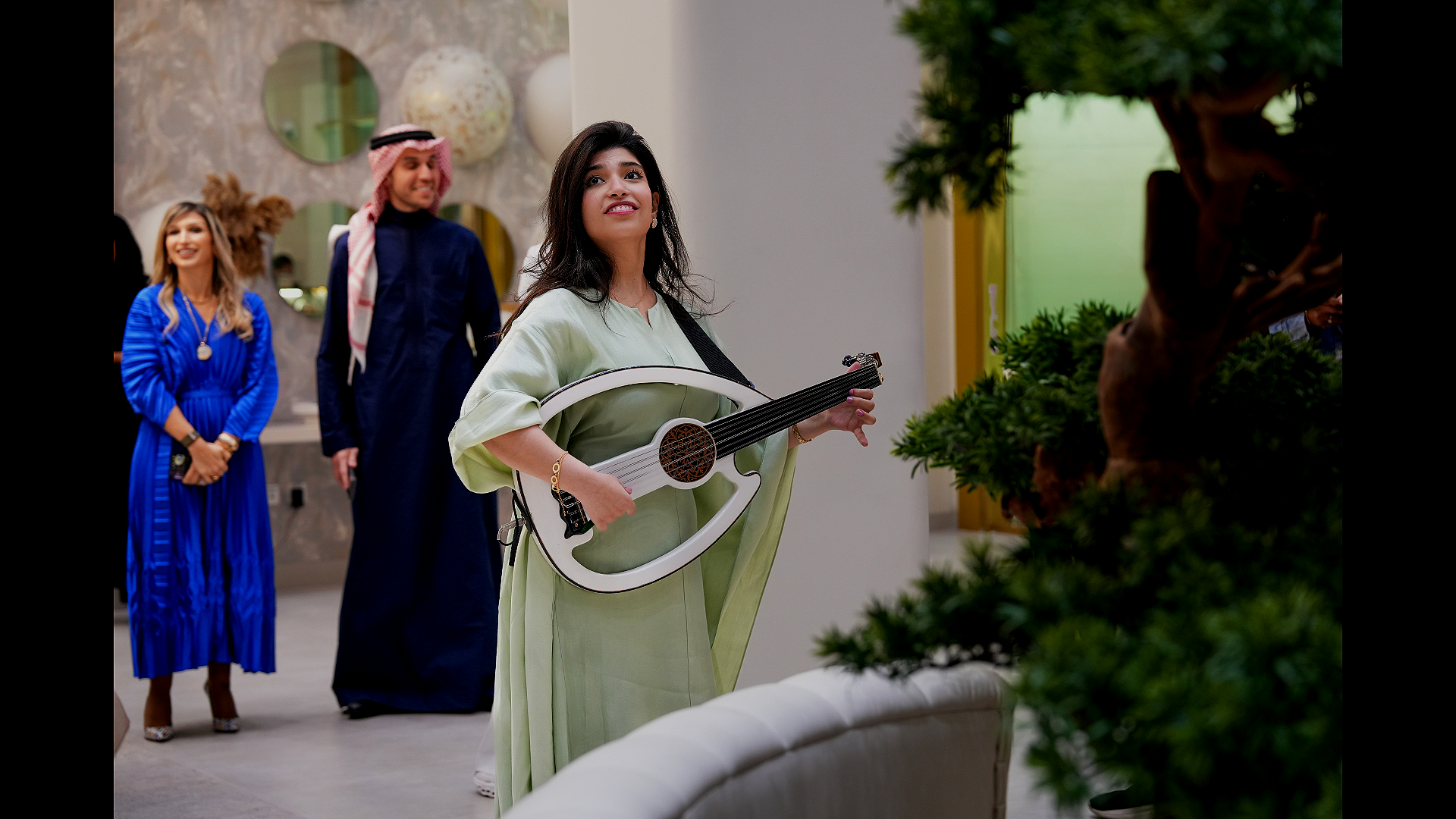A woman in a light green dress holding a white musical instrument, standing near a green plant, with three people in the background, one in a blue dress, another in traditional Middle Eastern attire, and a third in dark clothes, inside a modern building with decorative wall art.