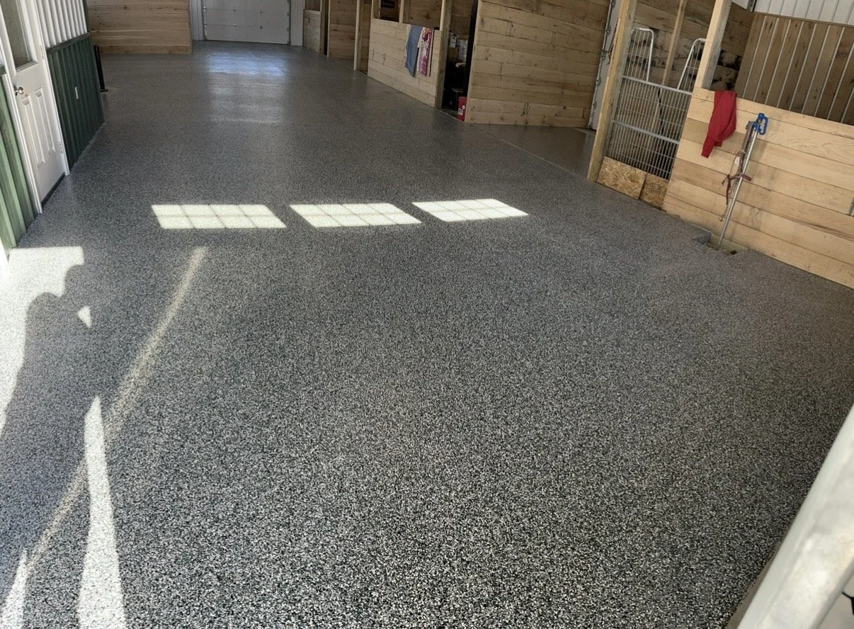 Interior of a barn or stable with a speckled epoxy floor, wooden walls, and metal stalls, illuminated by sunlight through windows.
