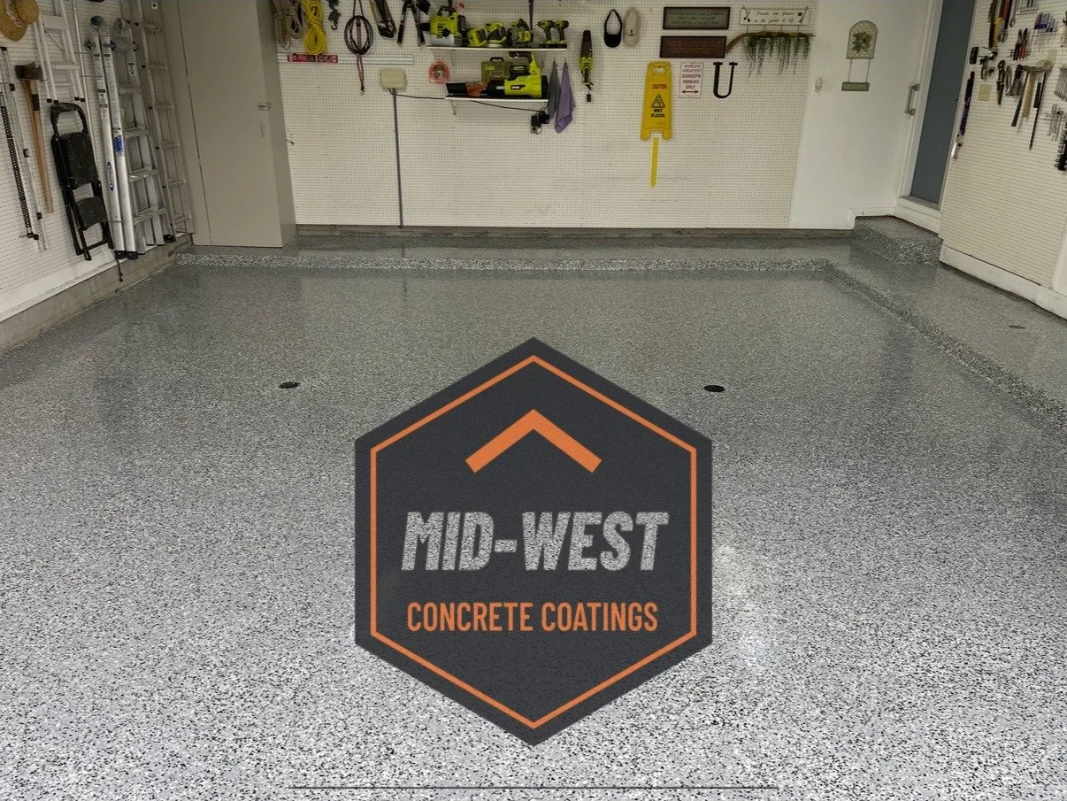 Empty garage with a newly coated concrete floor featuring a black and orange logo for Mid-West Concrete Coatings in the center. Shelves with tools and equipment are on the back wall, and a step is on the right side near a door.