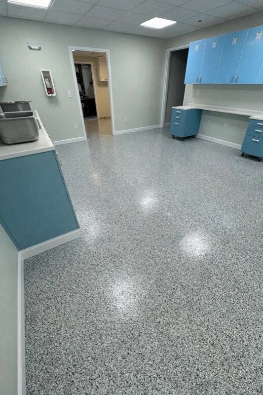 Empty medical or laboratory room with blue cabinets, counters, and stainless steel sinks, brightly lit with ceiling lights.