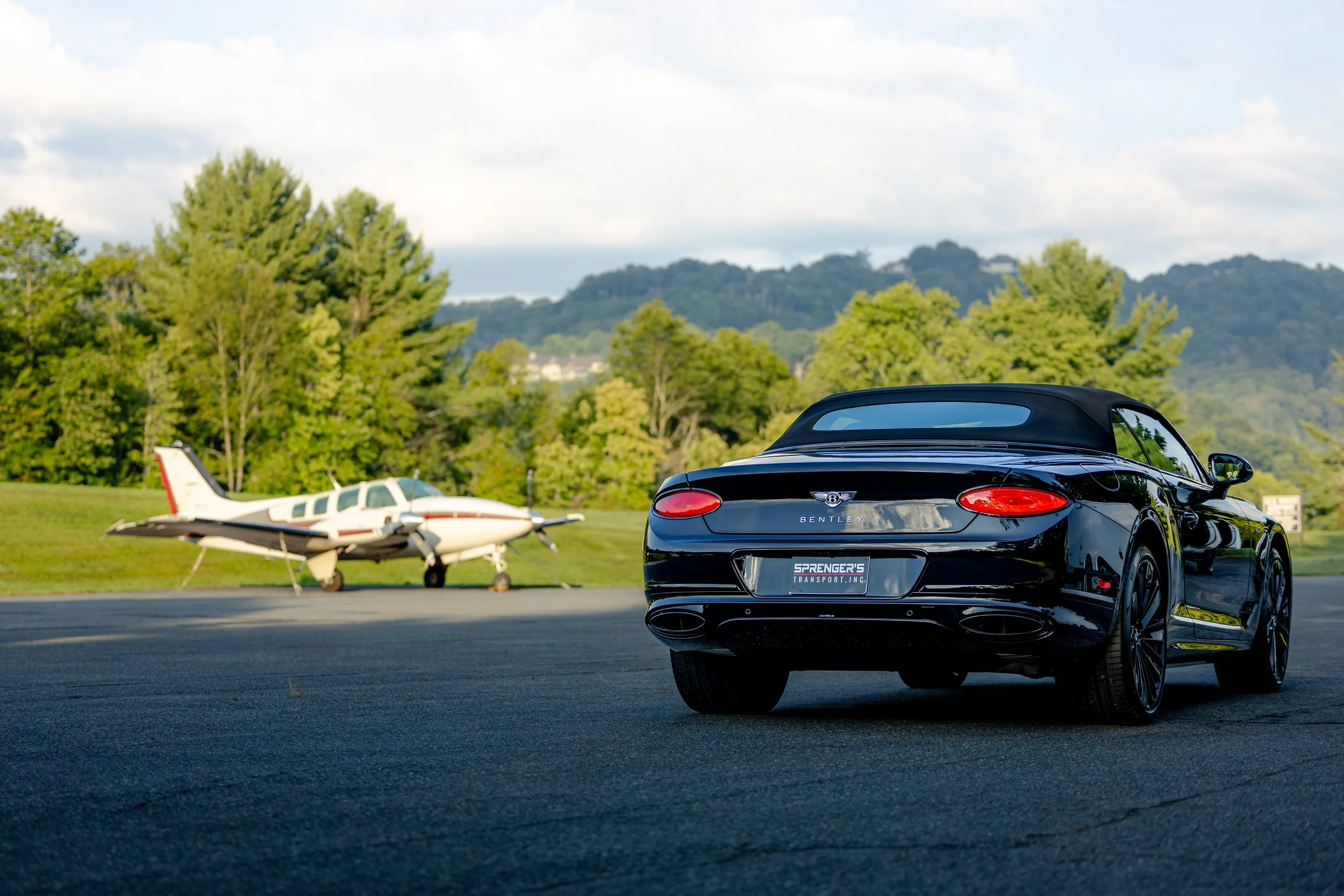 A sleek black Bentley Continental parked on an airstrip with small private planes and lush green trees and hills in the background.