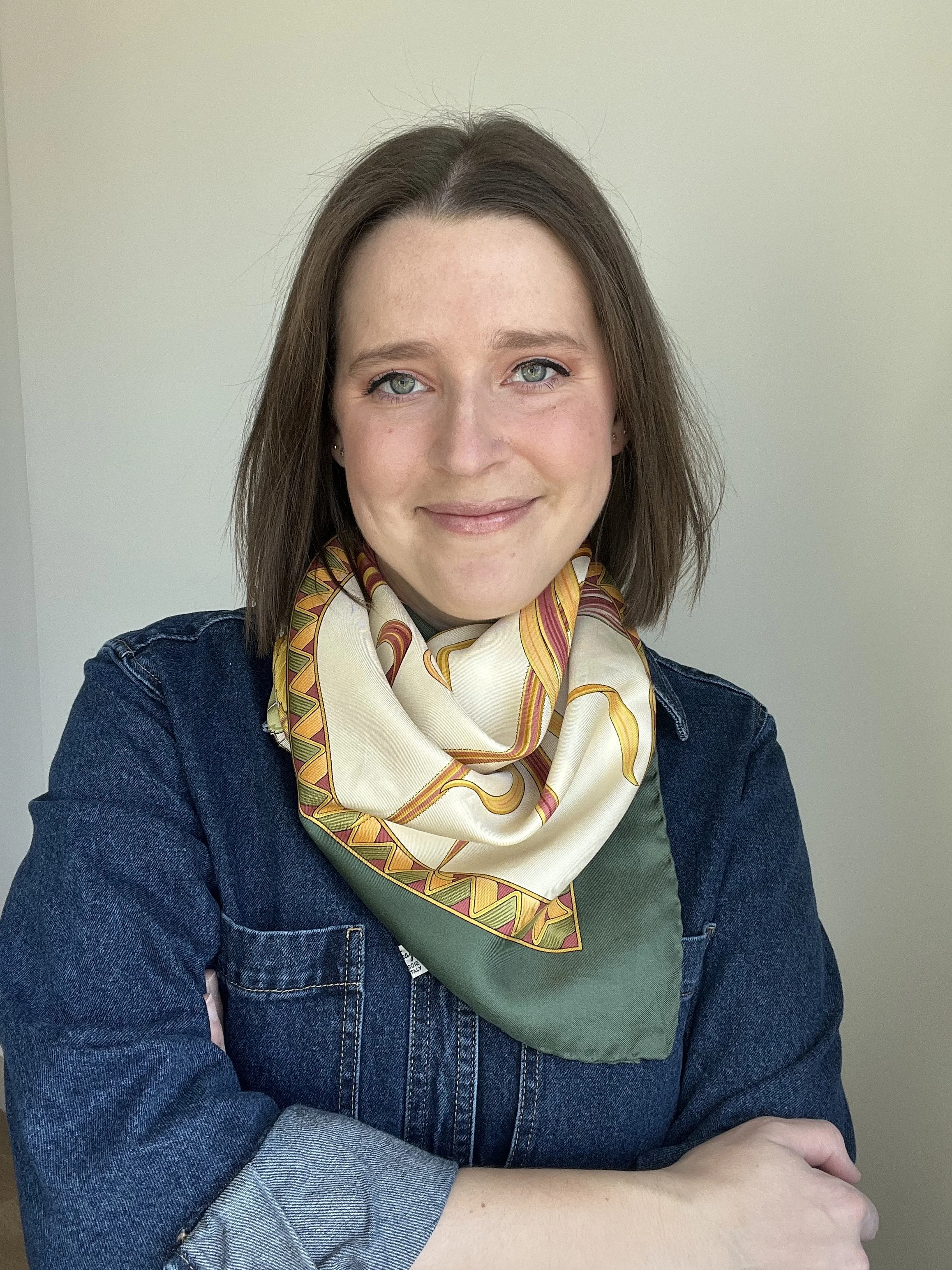 A woman with shoulder-length brown hair and green eyes smiling, wearing a denim jacket, olive green shirt, and a yellow patterned silk scarf, sitting against a plain light-colored background.