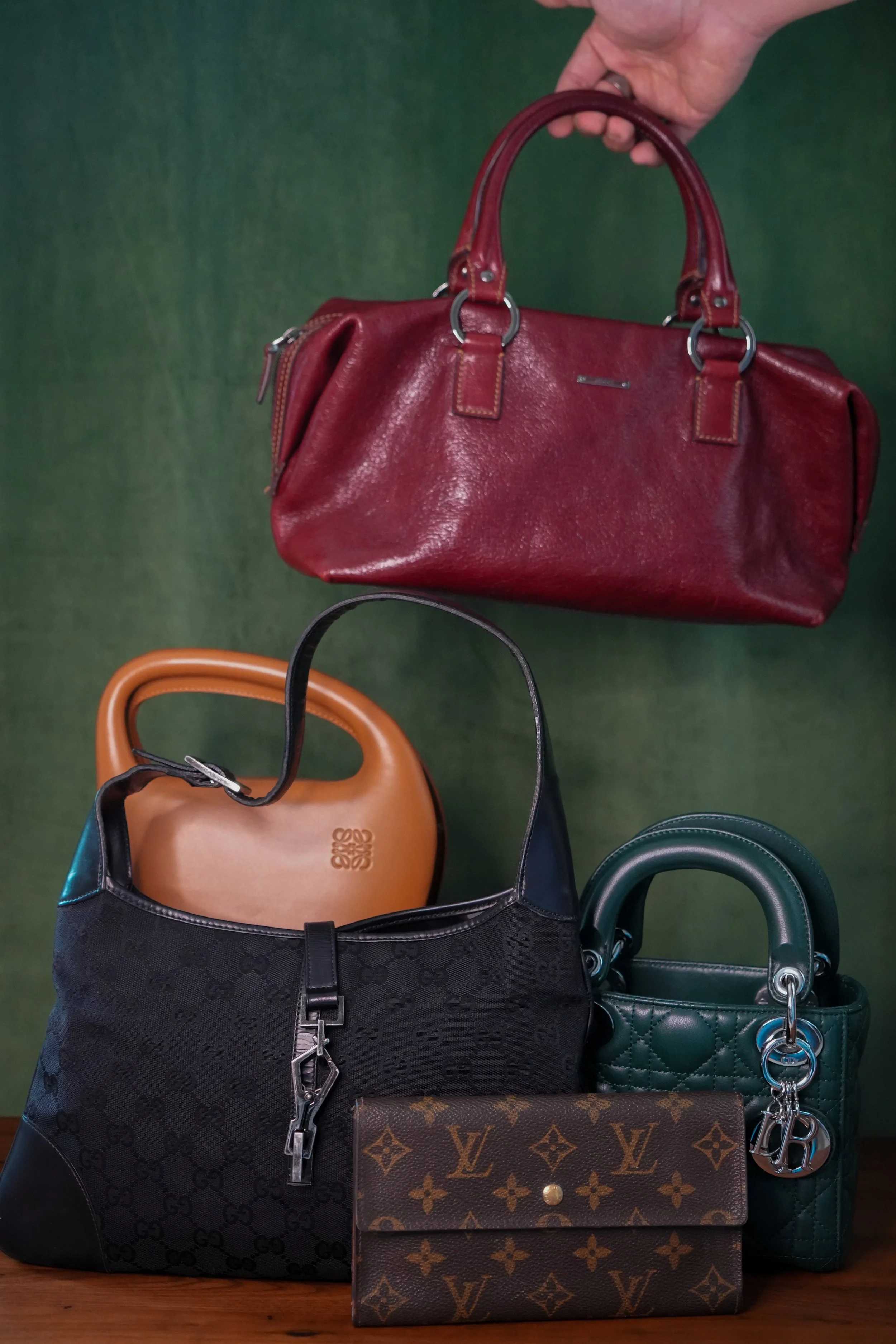 Various designer handbags and purses displayed on a table, including a red leather handbag, a black and gray patterned bag, a tan leather bag, and a Louis Vuitton wallet, with a green background.