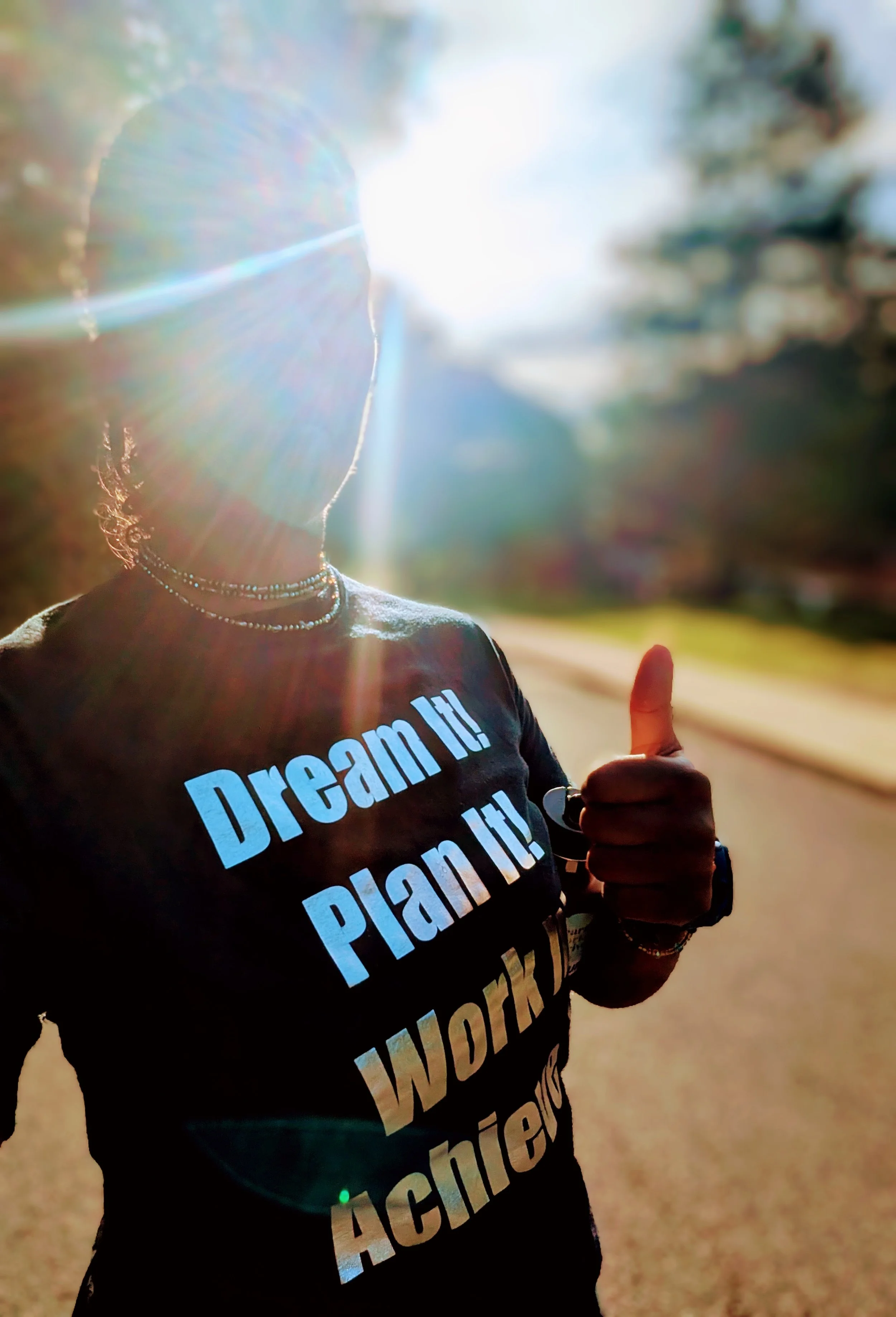 A woman wearing a black T-shirt with motivational text, multiple necklaces, and a watch, giving a thumbs-up gesture outdoors with the sun shining brightly in the background.