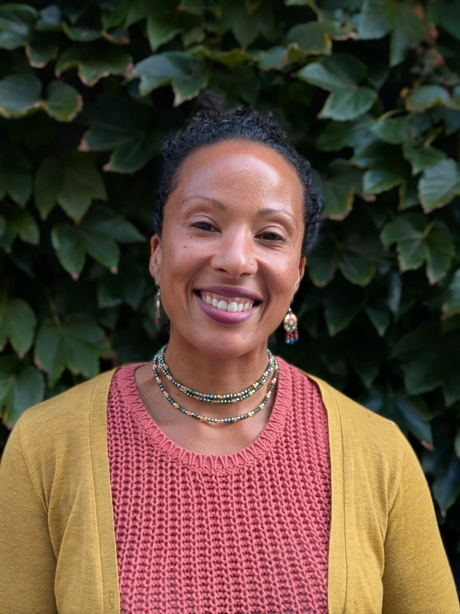 Close-up of a smiling African-American woman with natural hair pulled back, wearing a multiple necklaces and a yellow cardigan over a red knitted top, standing in front of green leafy bushes.