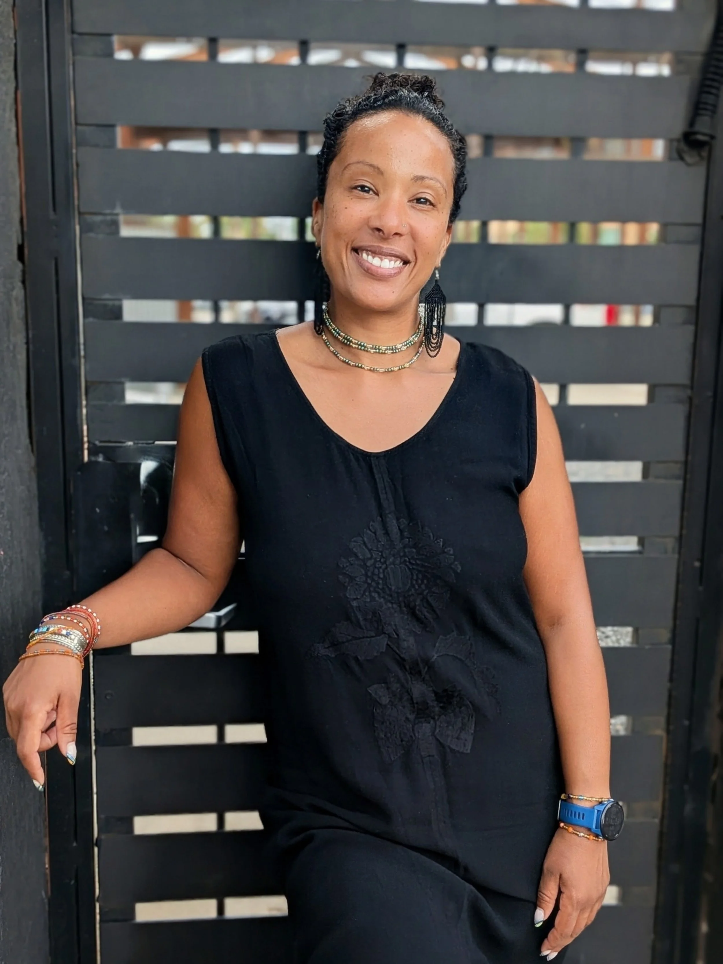 An African-American woman with curly hair tied back, wearing a black sleeveless dress, standing outdoors against a black slatted background, smiling warmly.