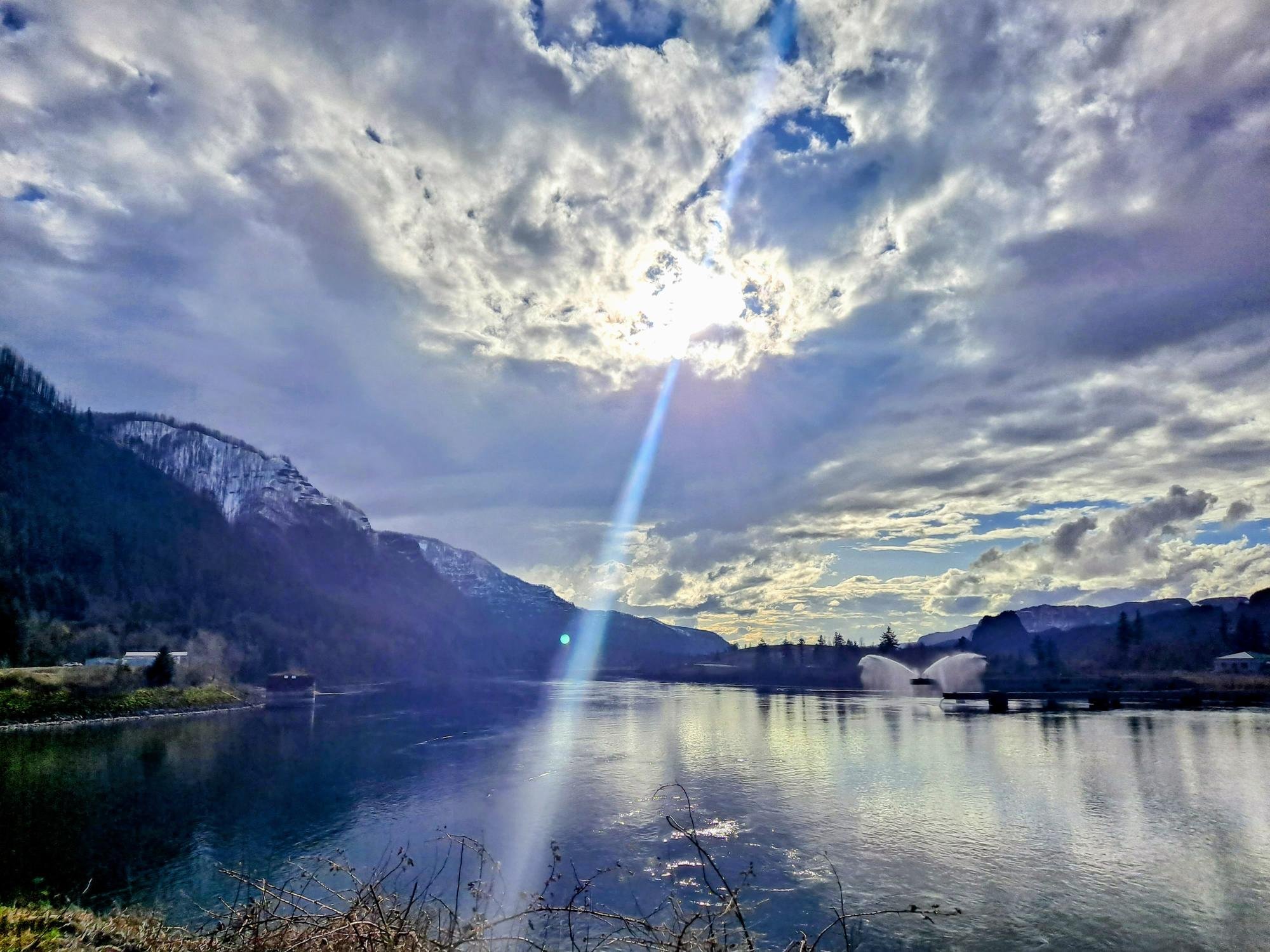 Sun shining through scattered clouds over a river with mountains in the background.