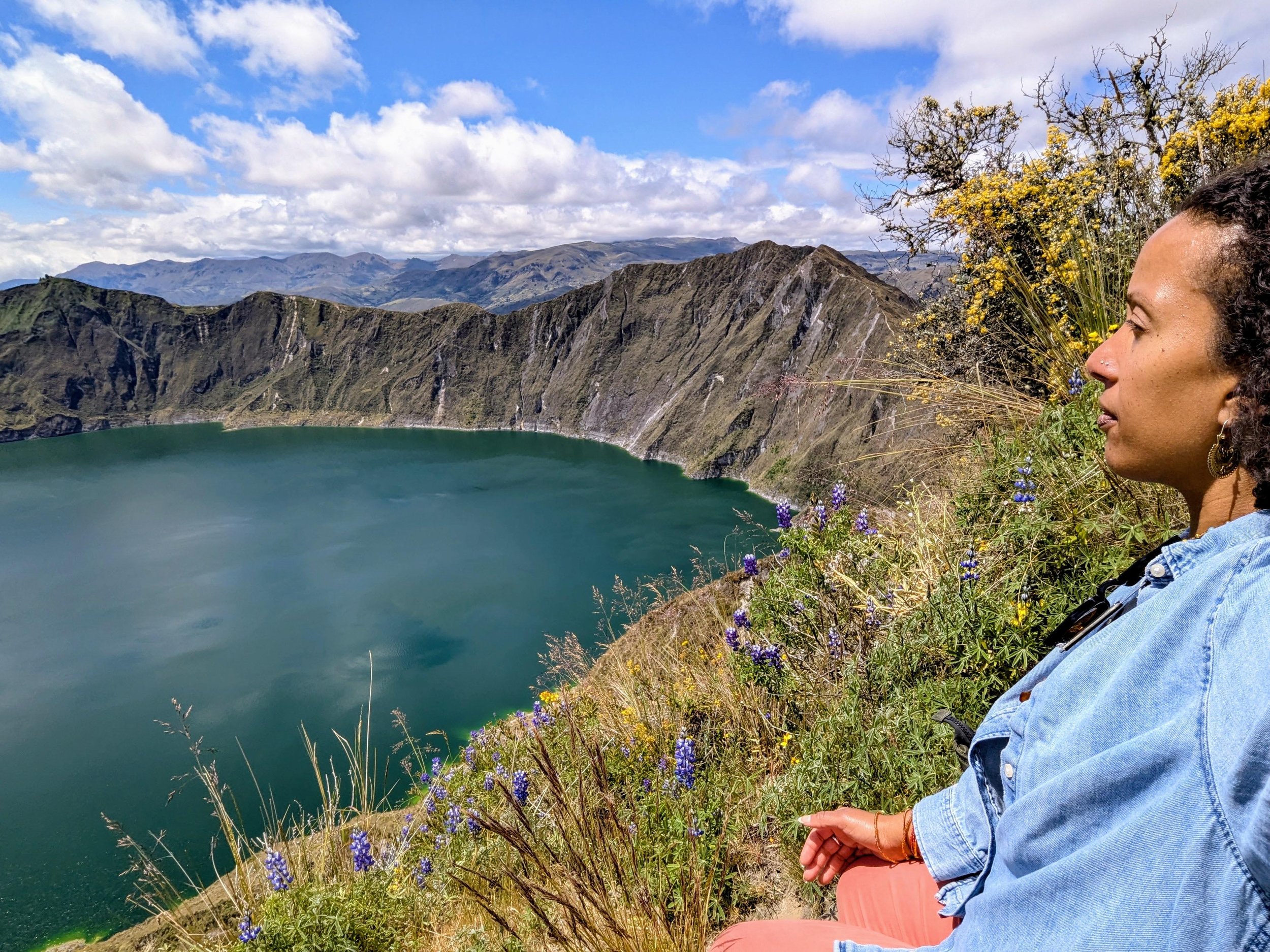 An African-American woman with curly hair and earrings is sitting on a hillside overlooking a volcanic crater lake in Ecuador, with mountains and a partly cloudy sky in the background.