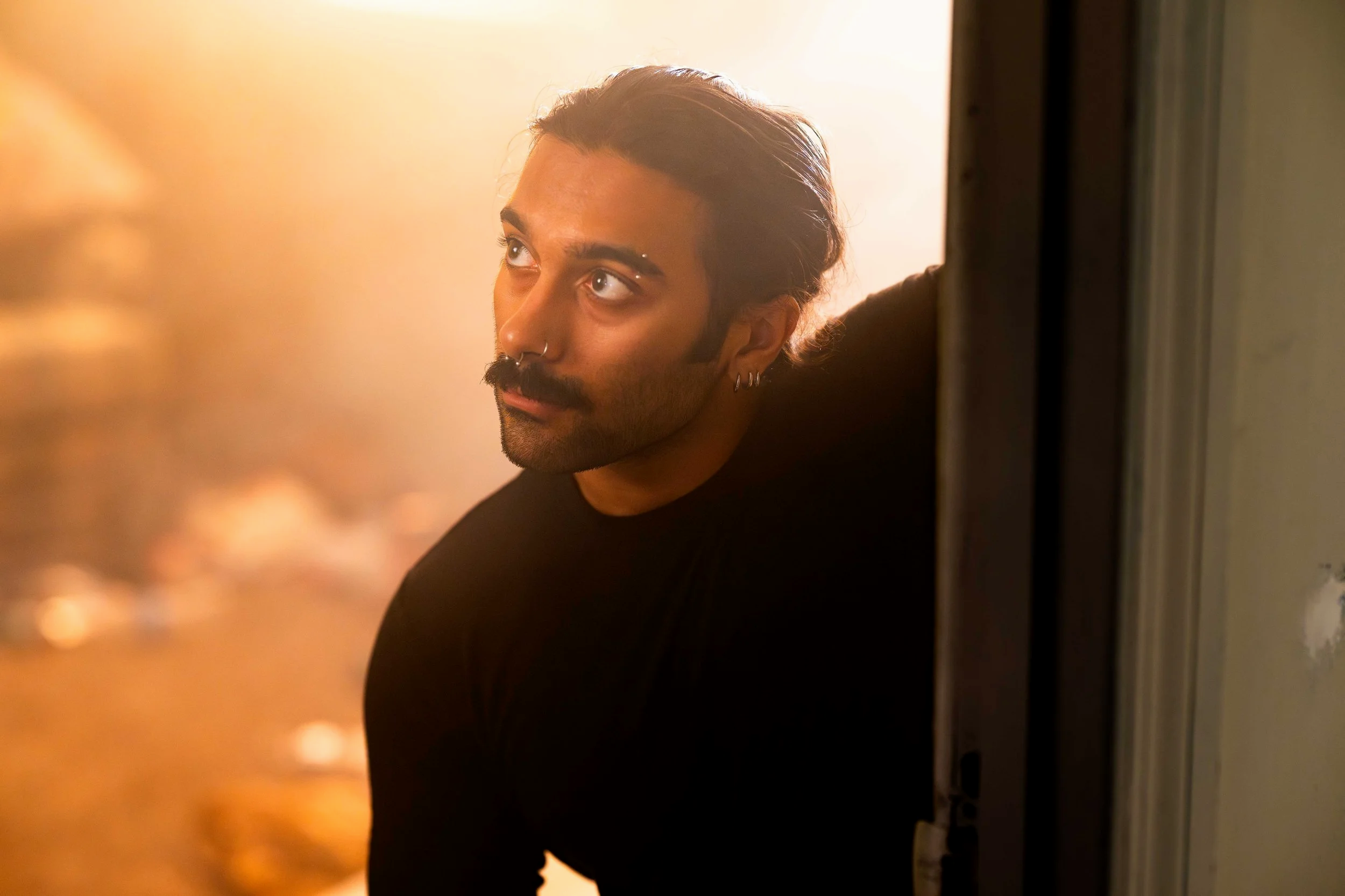A young man with dark hair and facial hair, wearing earrings and a nose ring, peeking from behind a wall or door, looking to the upper left with a thoughtful expression. The background is warm and softly lit.