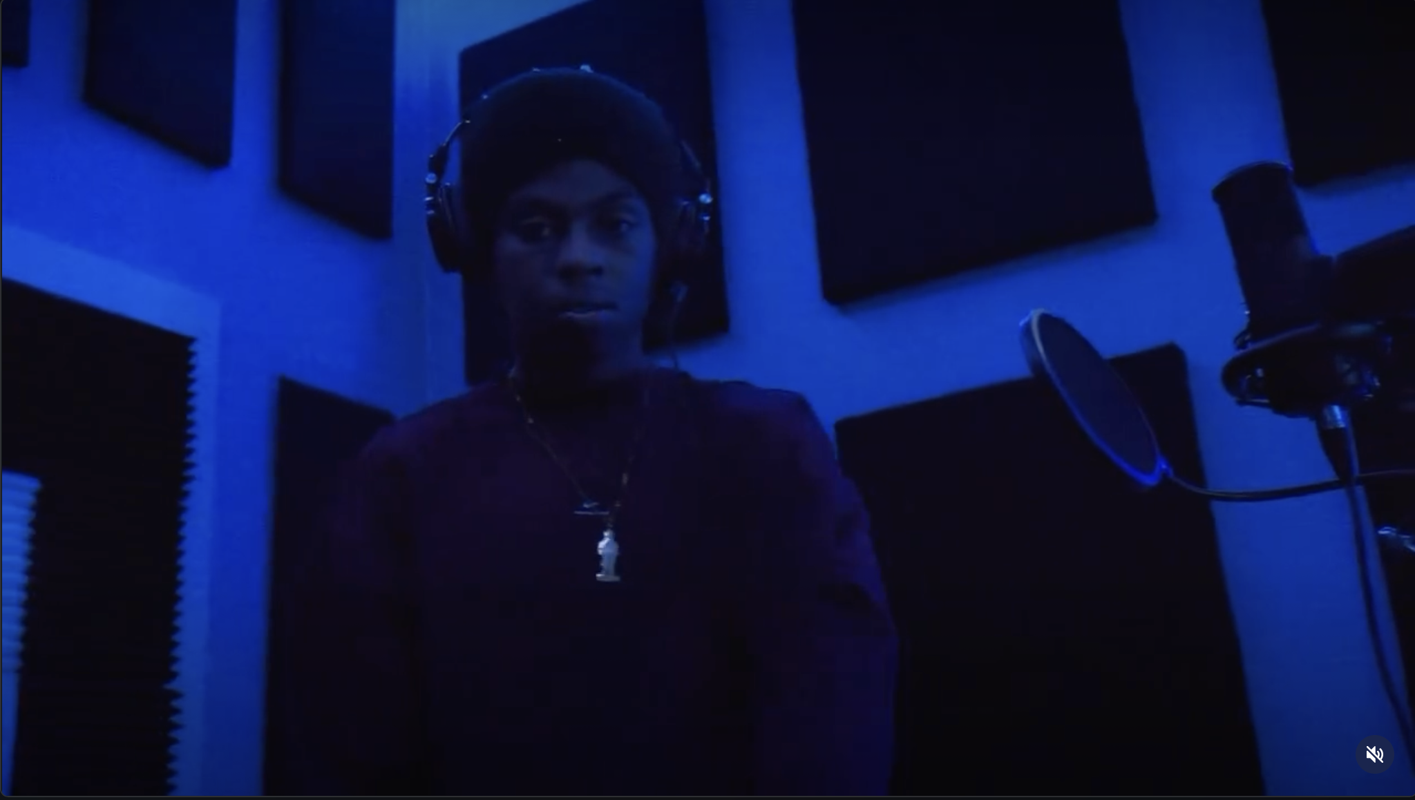 A person in a dark room wearing headphones and a beanie, standing in front of a microphone in a music recording studio, with blue lighting and soundproofing panels on the walls.