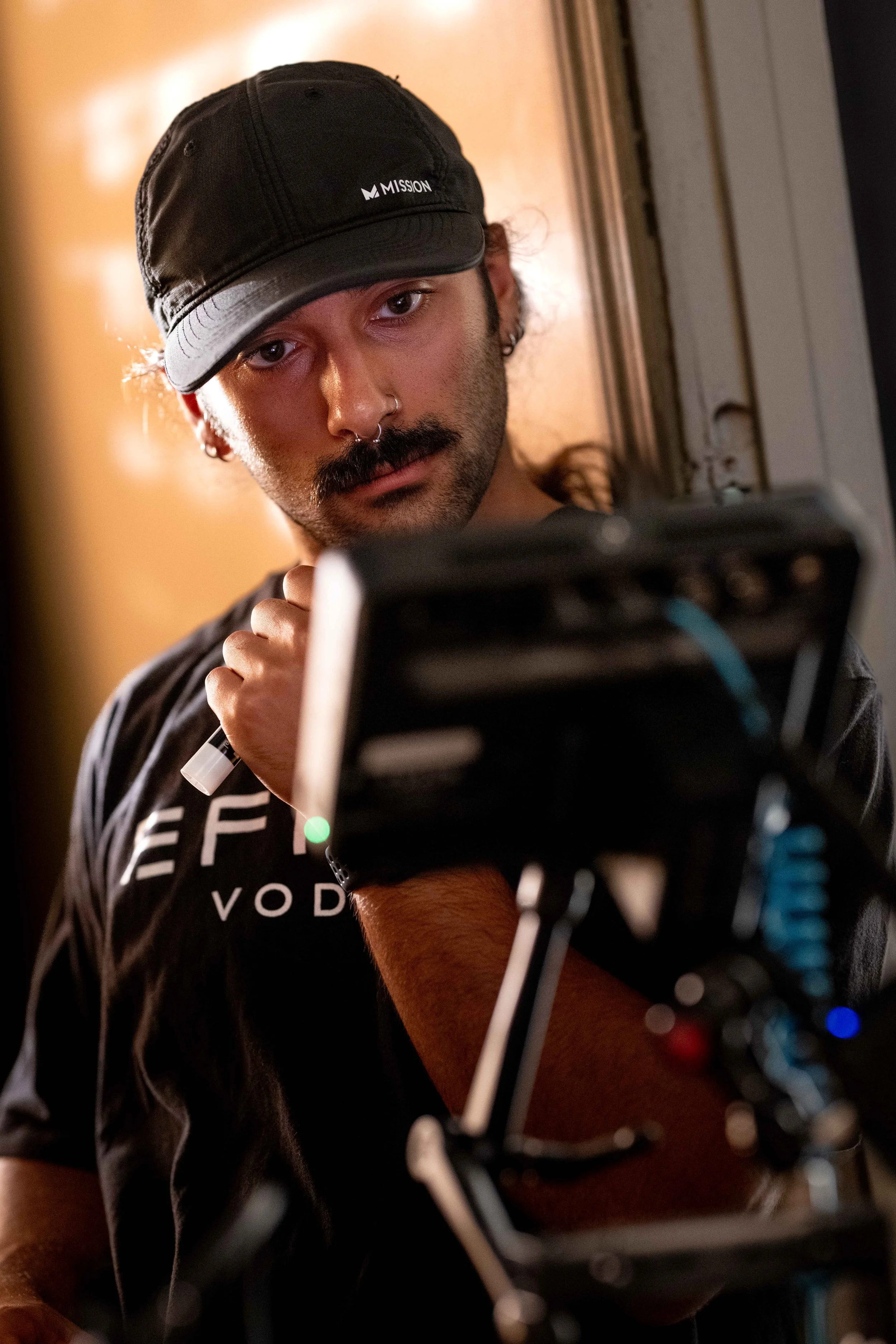 A man with a black cap, nose ring, and mustache working on sound equipment, possibly a mixer or audio control panel, in an indoor setting.