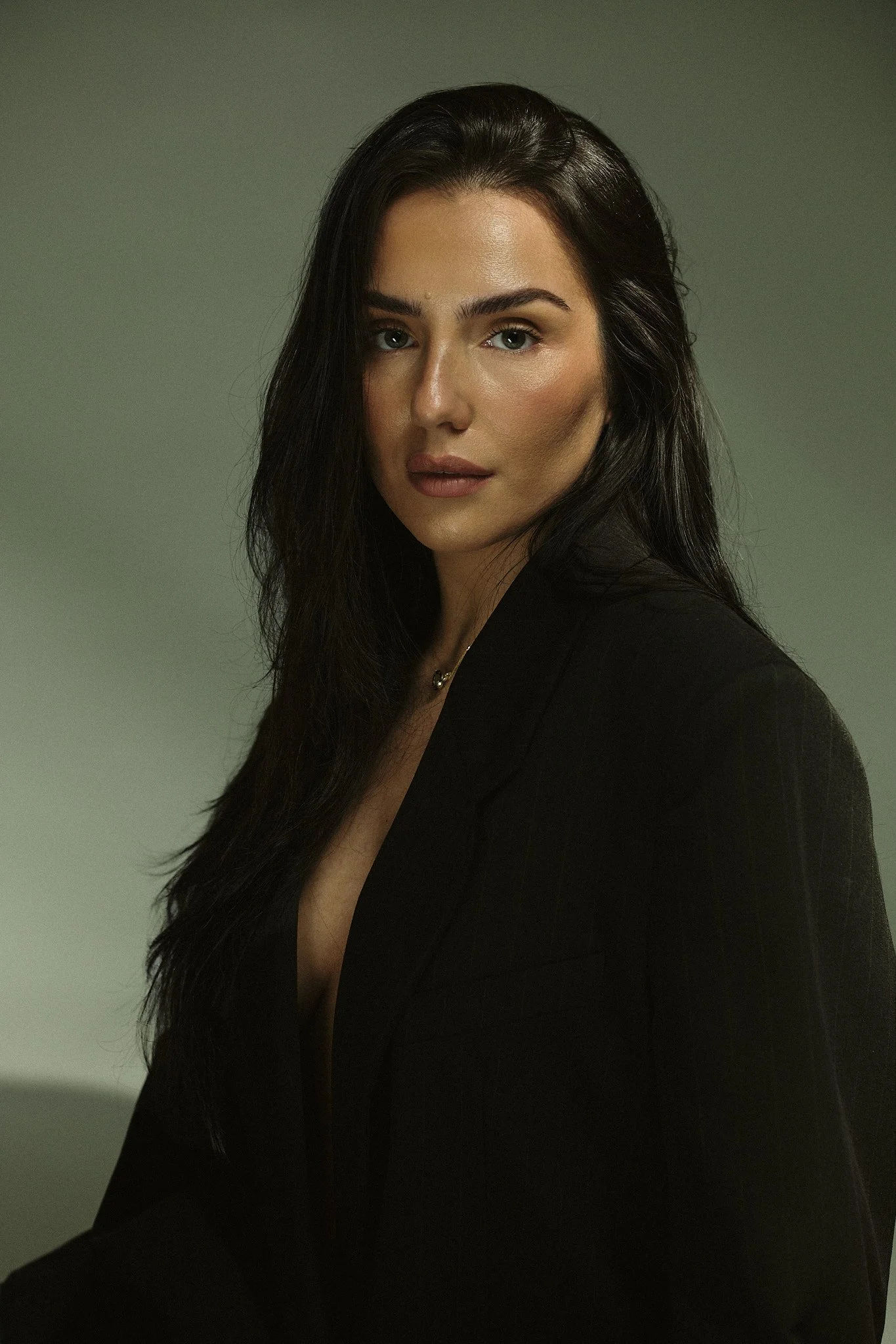 A woman with long dark hair, wearing a black blazer, gazes at the camera against a plain gradient background.