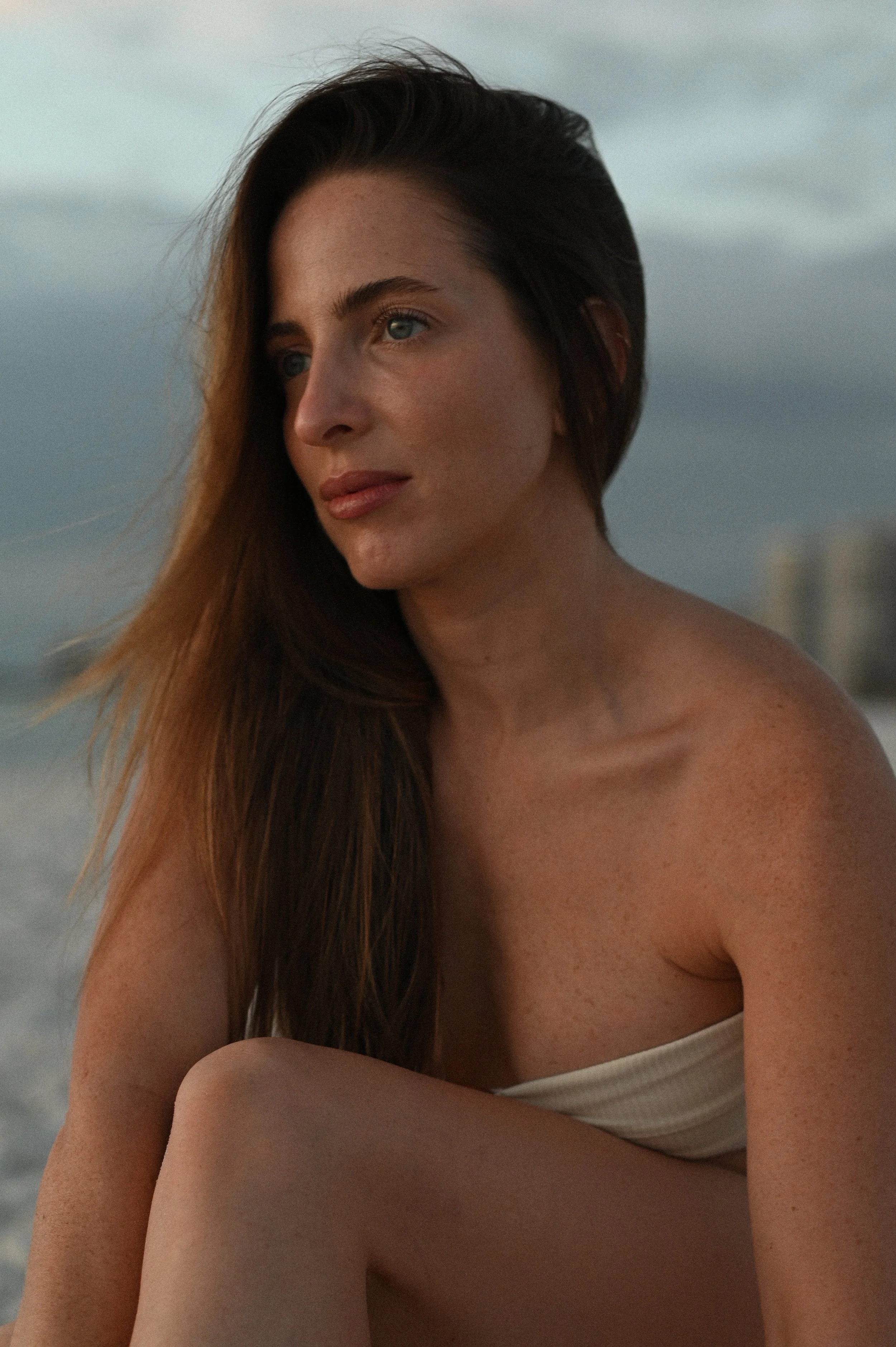 A young woman with long brown hair and blue eyes looking thoughtfully into the distance at a beach with a cloudy sky.