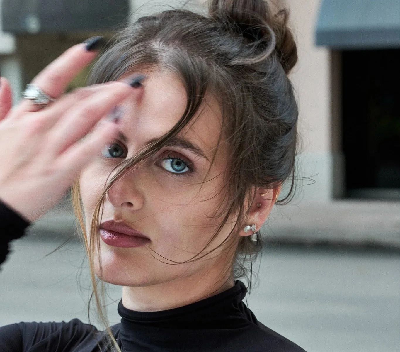 Close-up of a young woman with blue eyes and brown hair pulled up, touching her face with her right hand, wearing earrings and a black top.